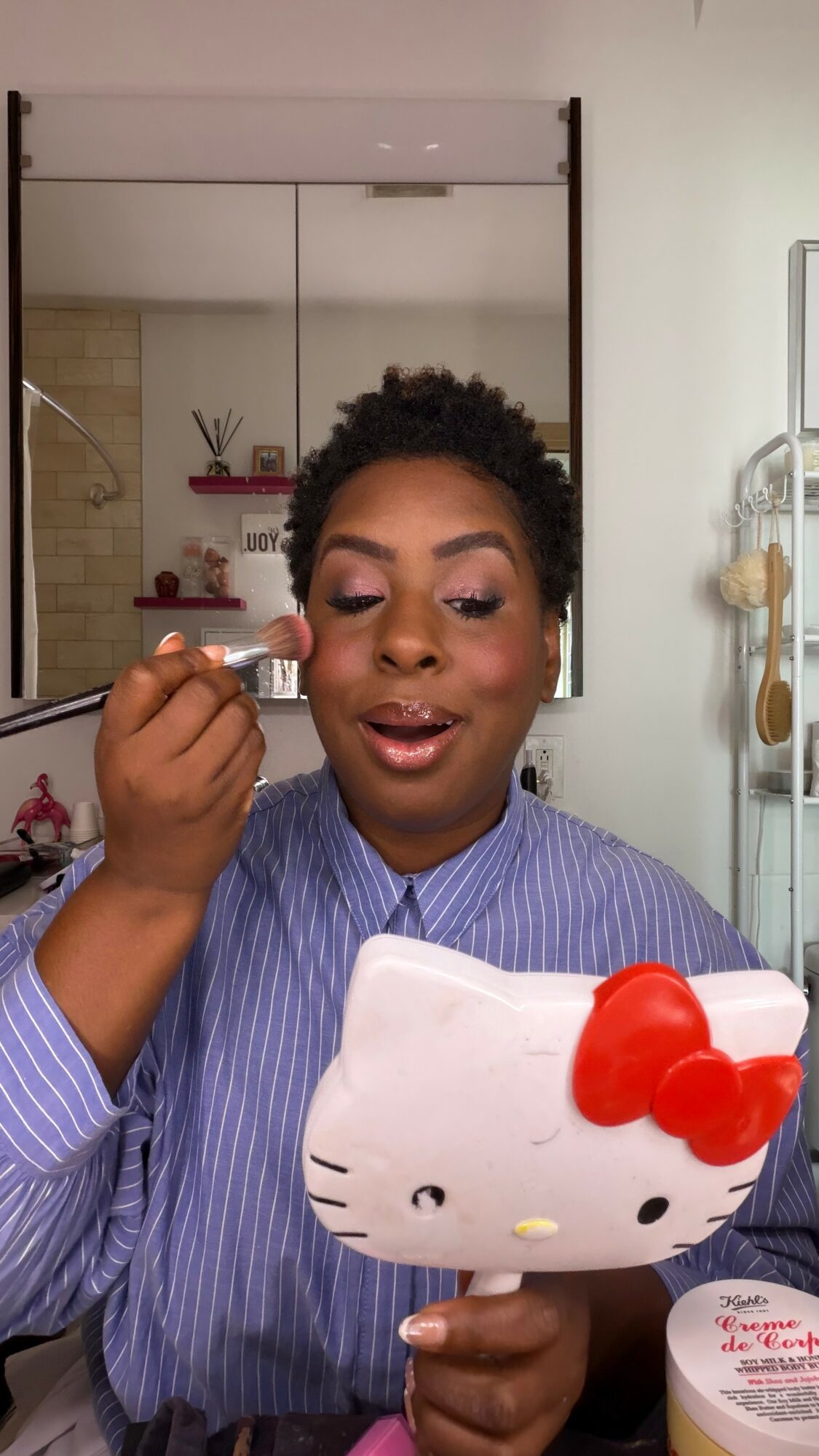 Woman with short curly hair applying makeup, holding a Hello Kitty mirror, in a room with shelves and mirror behind her.