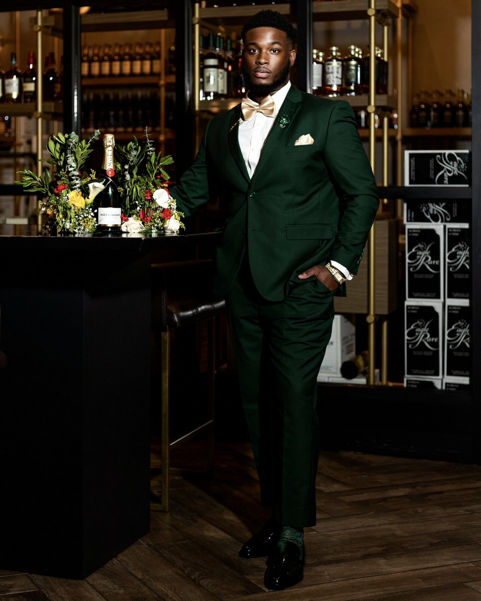 Man in green suit with bow tie standing in a bar with flowers and bottles behind him.