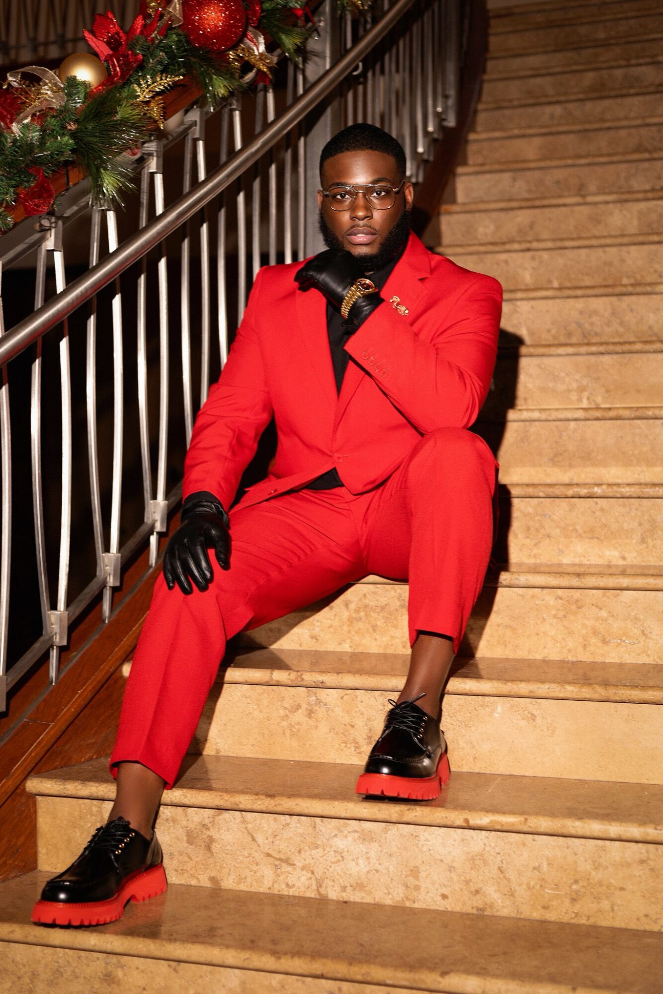 Person in red suit sitting on staircase, wearing black gloves and shoes, with a festive decoration nearby.