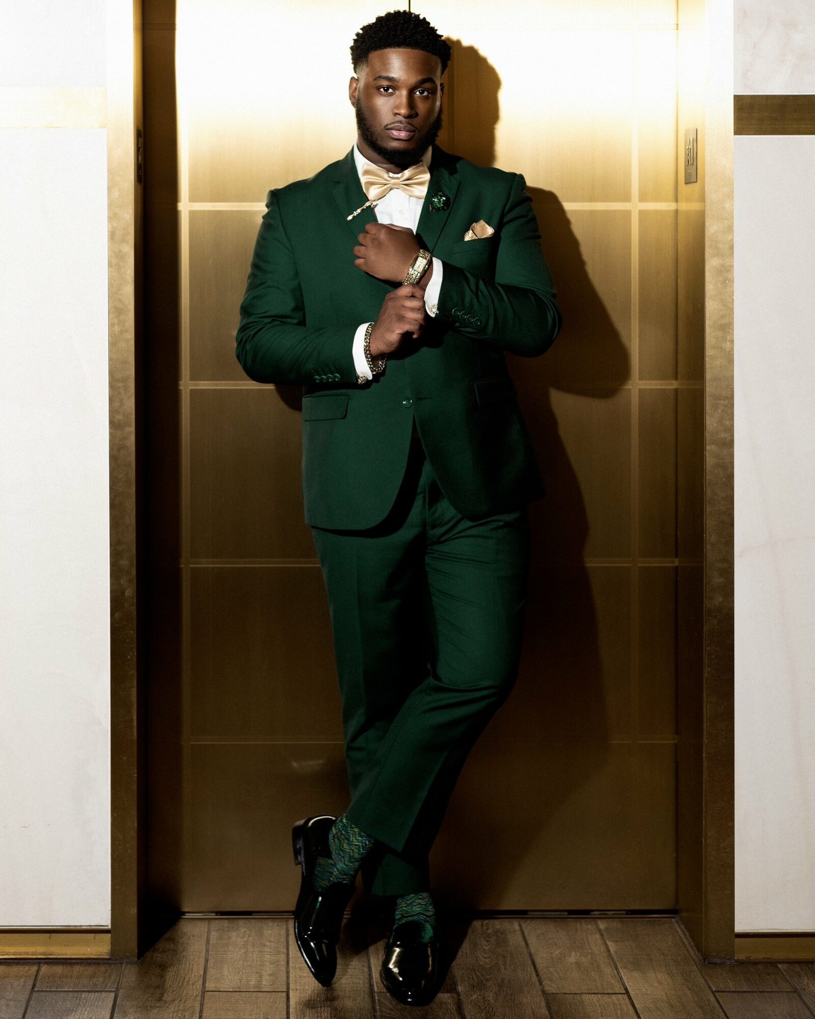Man in a green suit with a bow tie standing in an elevator, arms crossed, looking at the camera.