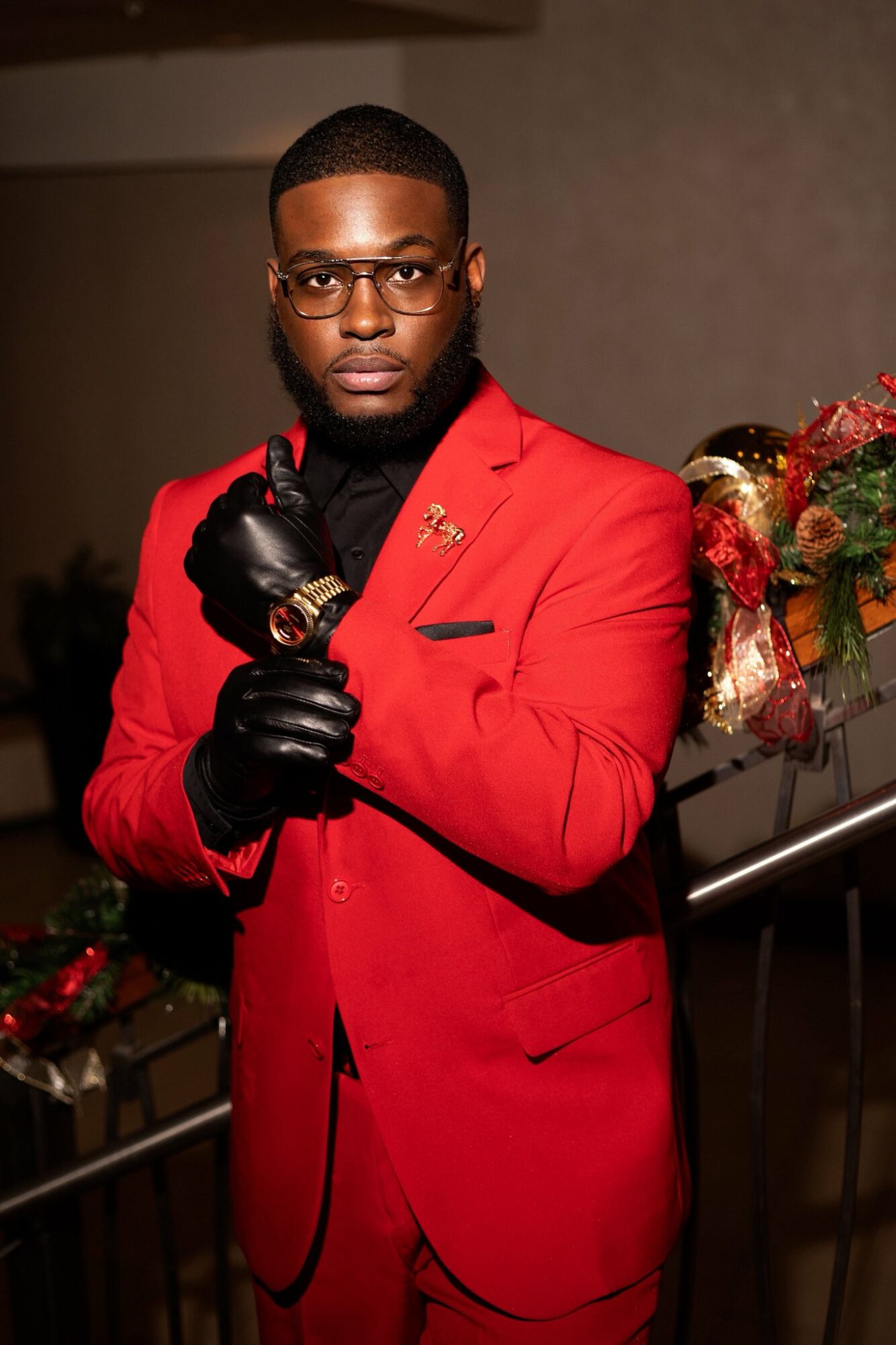 Man in red suit with black gloves and glasses, standing near a decorated Christmas tree, holding a cigar, indoors.
