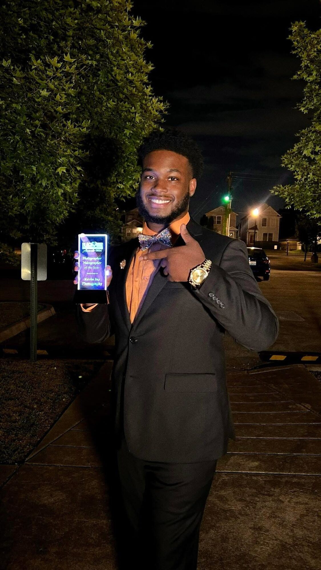 Man in suit holding a smartphone outdoors at night, smiling, pointing at the phone, trees and streetlights in background.