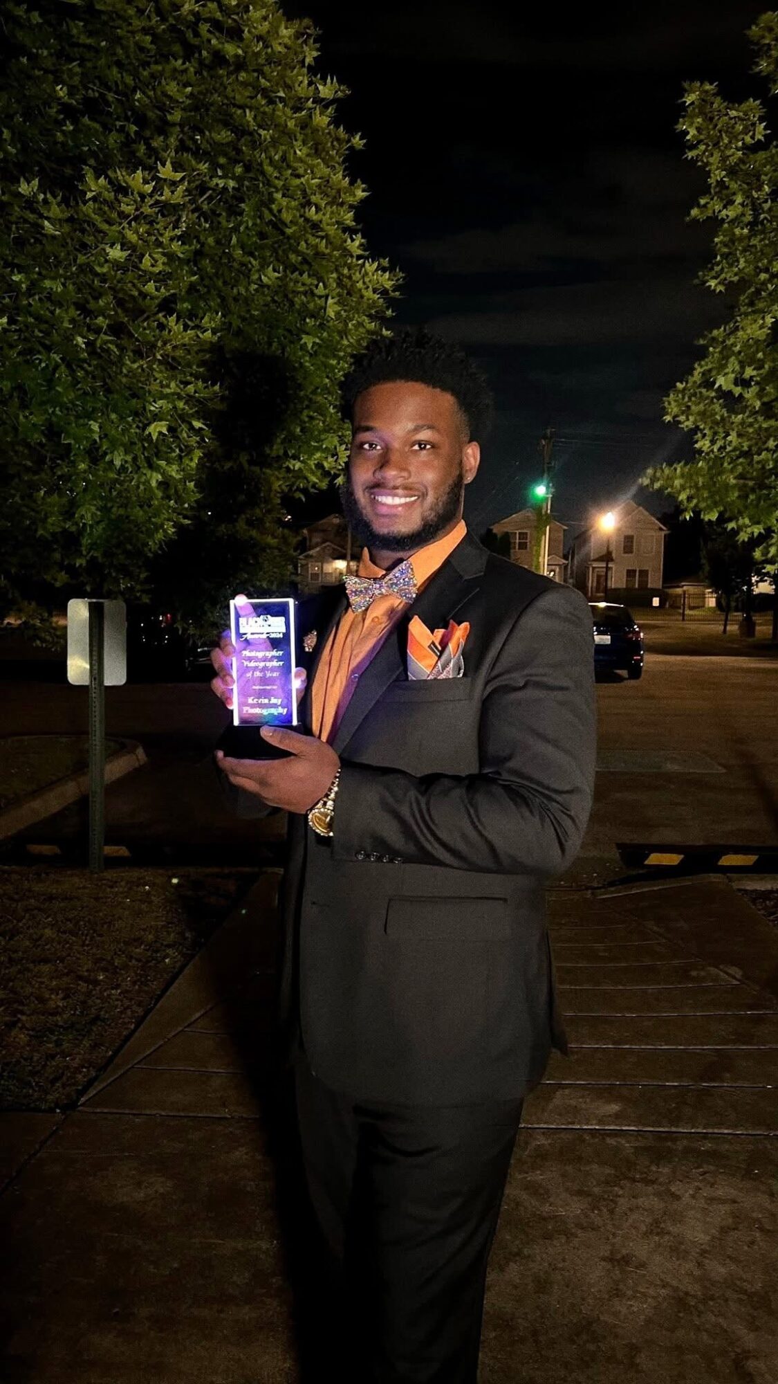 Man in suit holding a phone outdoors at night, smiling, trees and streetlights in background.
