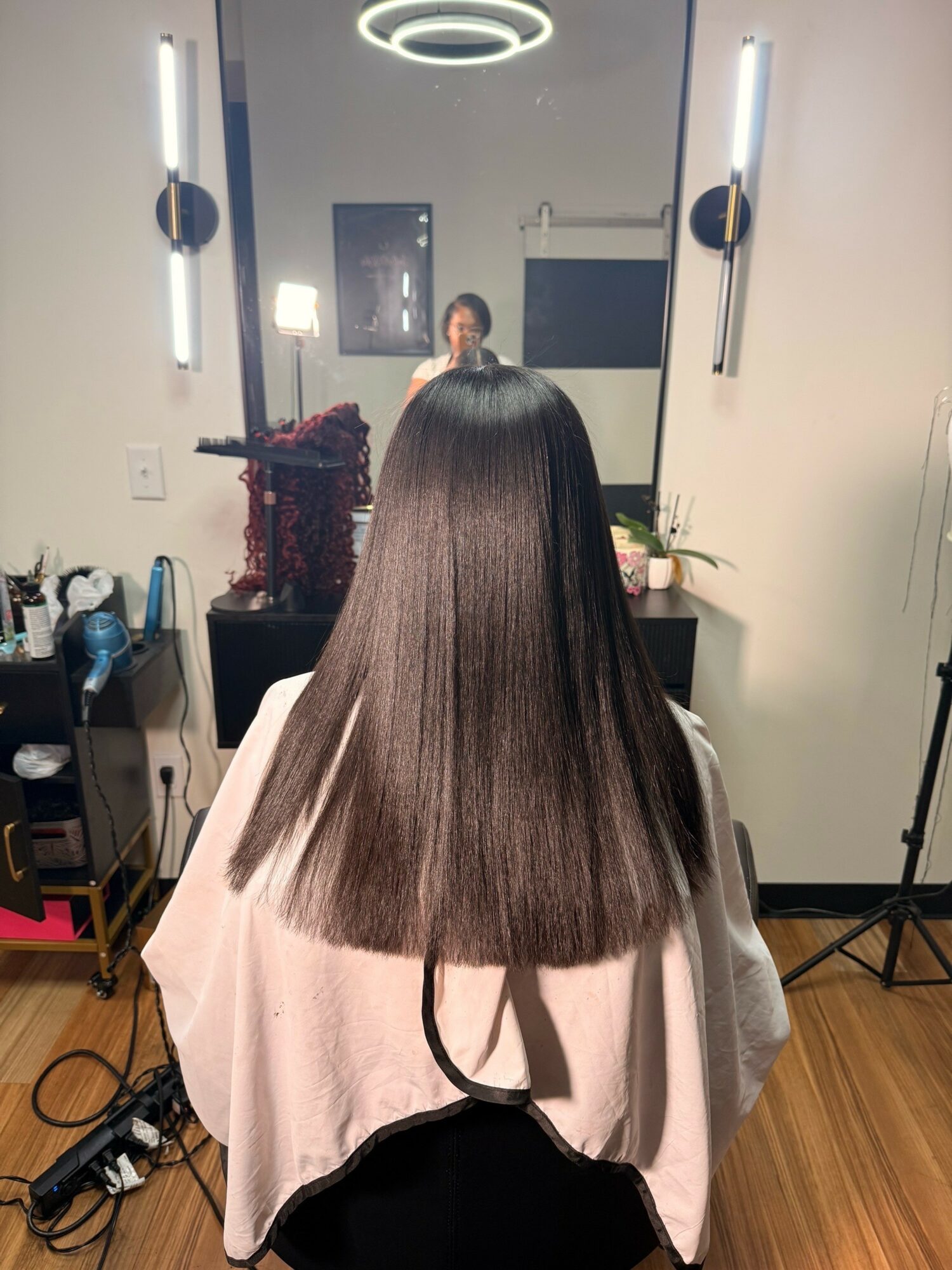 Person with straight, shiny hair sitting in a salon chair, facing a mirror with a person taking a photo.
