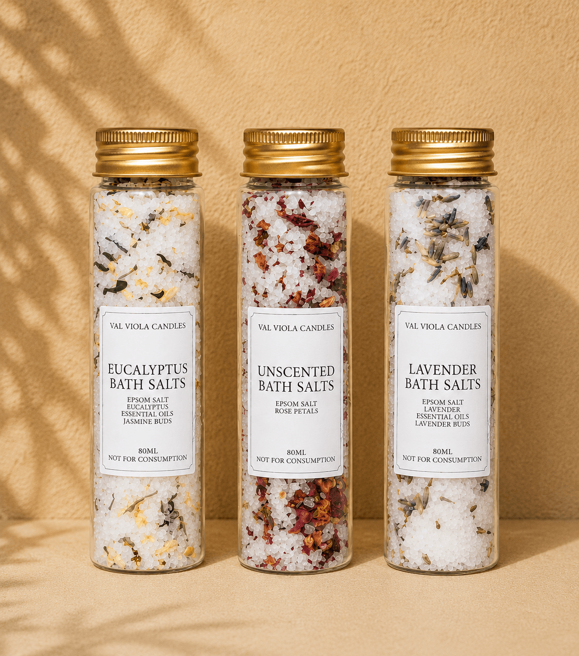Three glass jars with gold lids containing bath salts, labeled eucalyptus, unscented, and lavender, against a beige background.