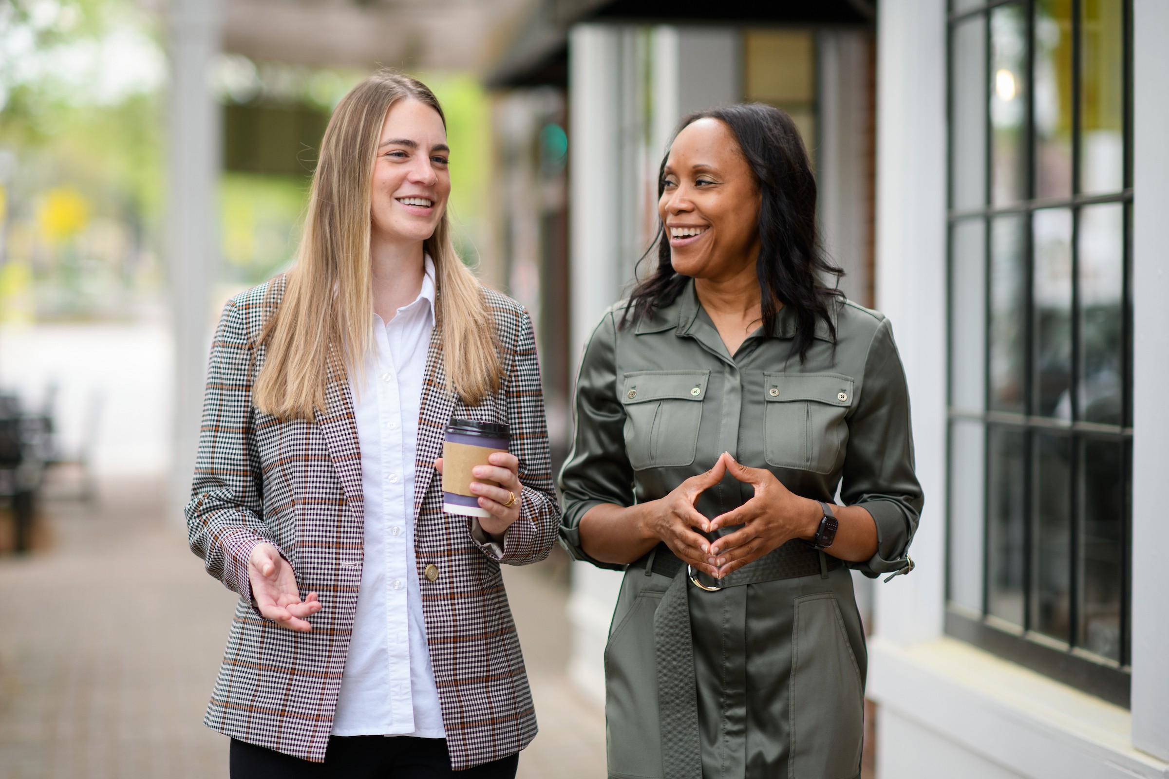 Two women are walking and talking outside, smiling and holding coffee cups, near a building with large windows.