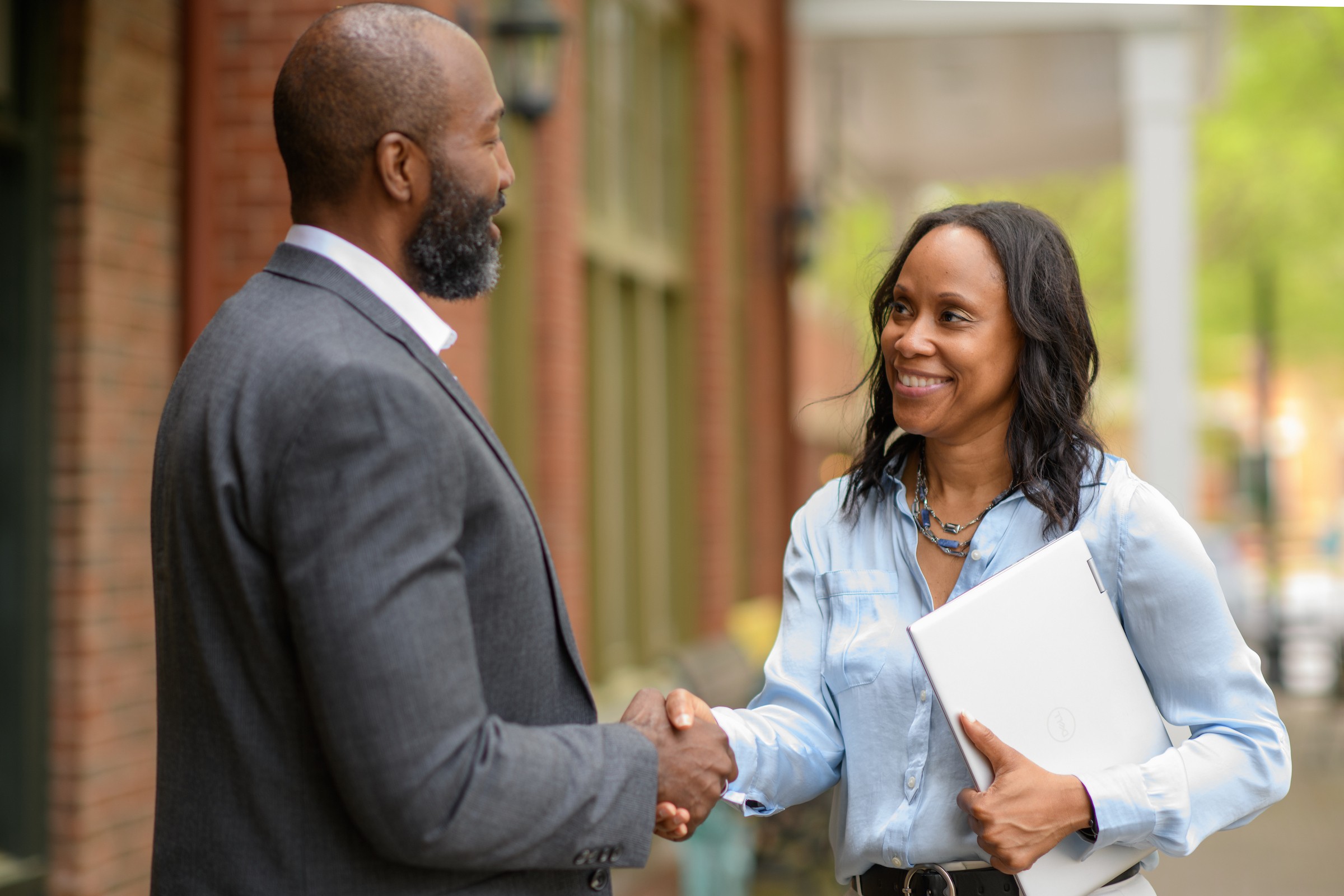 Two people shaking hands outdoors, one holding a folder, smiling, with a blurred background of buildings and trees.