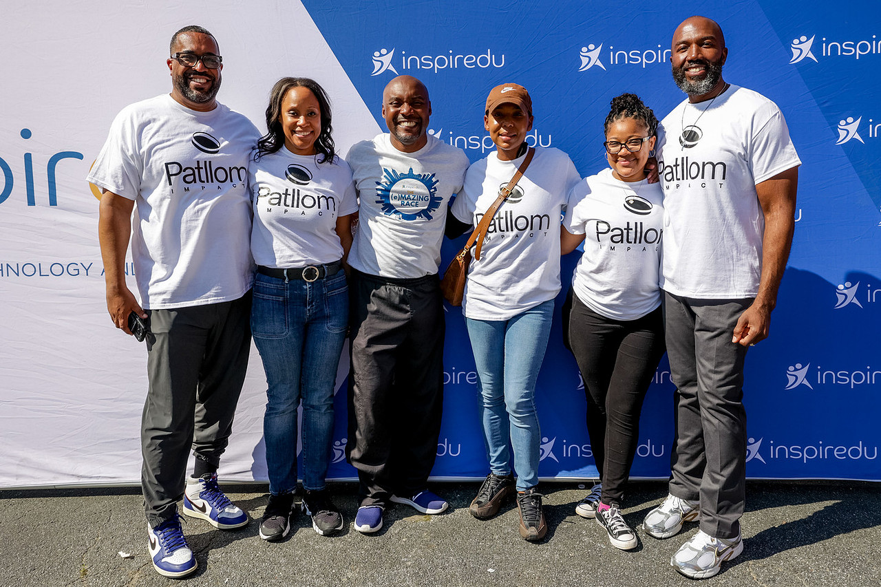 Group of six people standing outdoors in front of a blue and white backdrop, smiling, wearing casual clothes and white t-shirts.