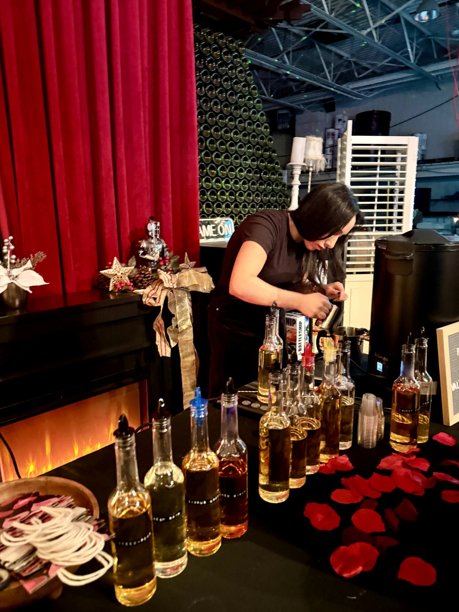 A woman prepares drinks behind a bar with bottles, red roses, and a fireplace, in a decorated indoor setting.