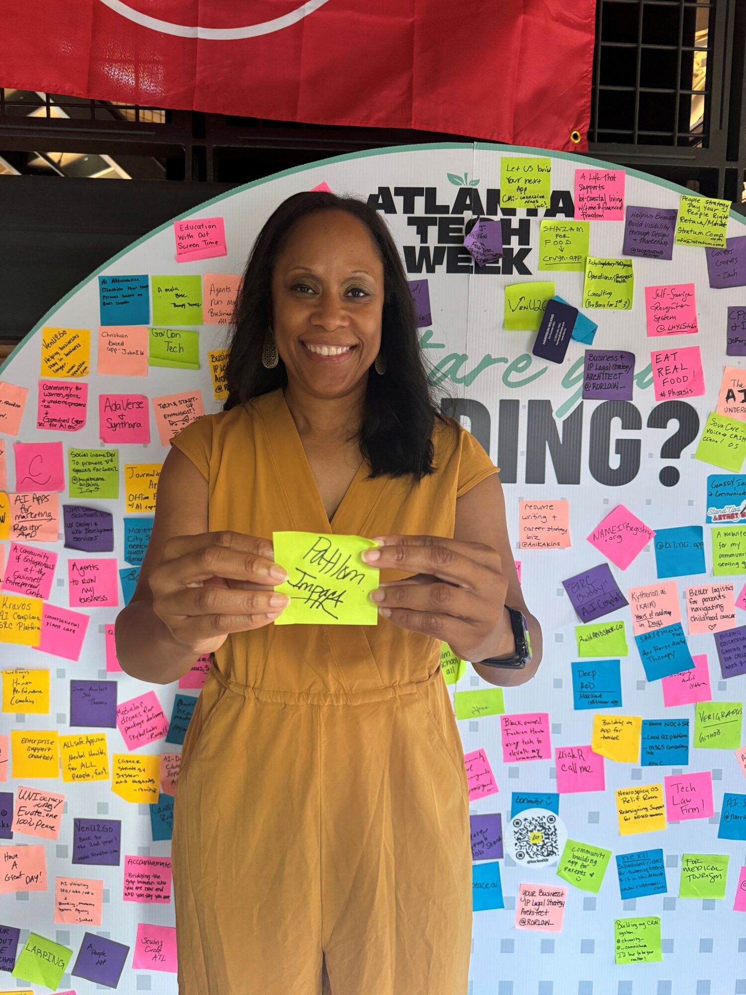 Woman in yellow dress holding a yellow sticky note, standing in front of a colorful sticky note display at an event.