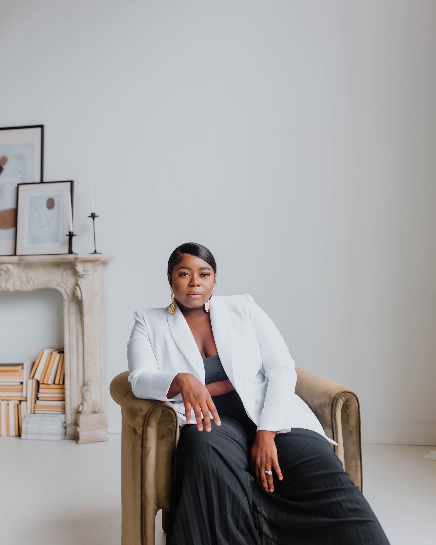 Woman sitting in a beige armchair in a minimalistic room with a white wall and a fireplace with books and framed pictures.