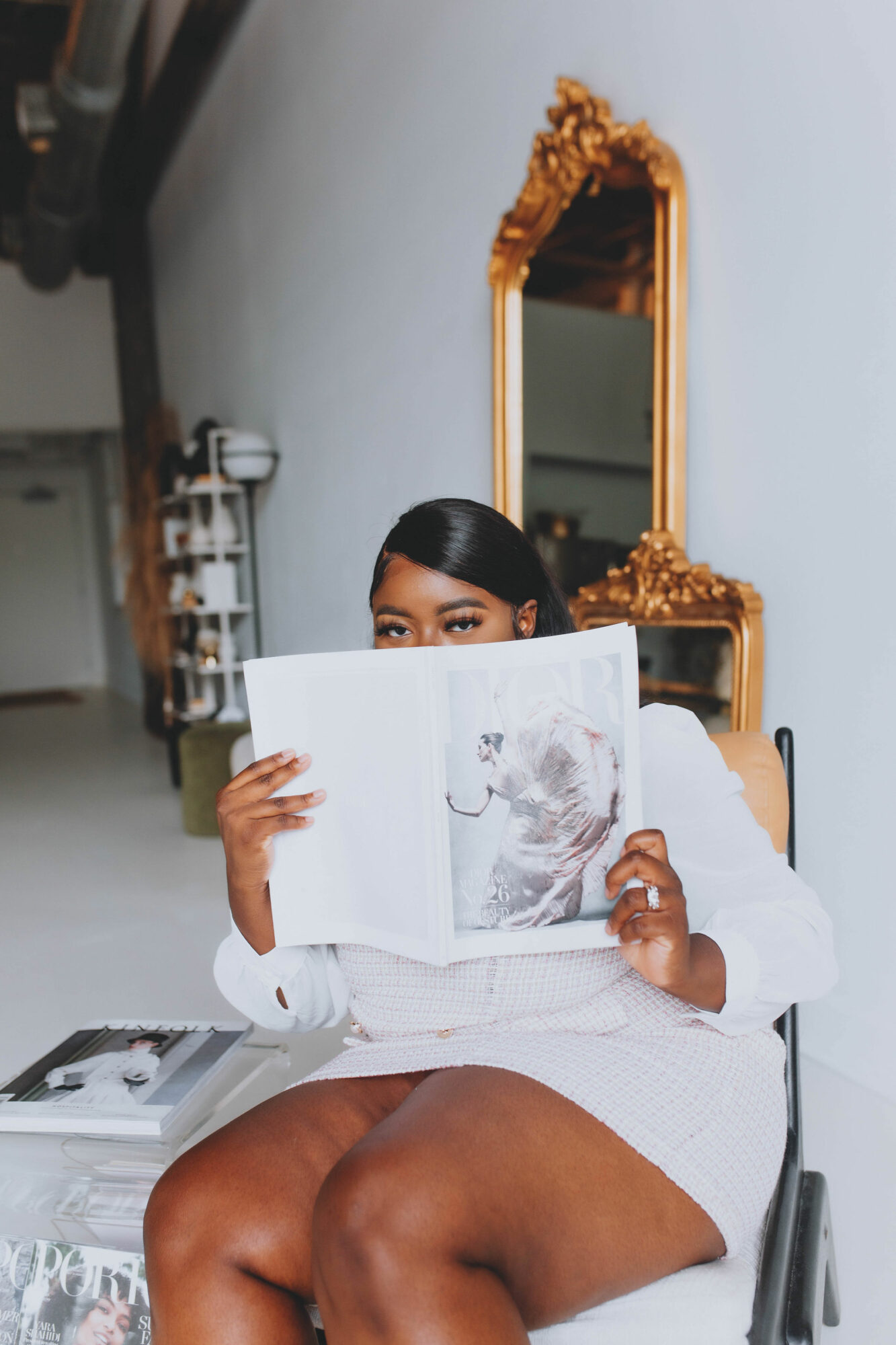 Person sitting on a chair holding an open magazine, with a mirror and decorative mirror frame in the background.
