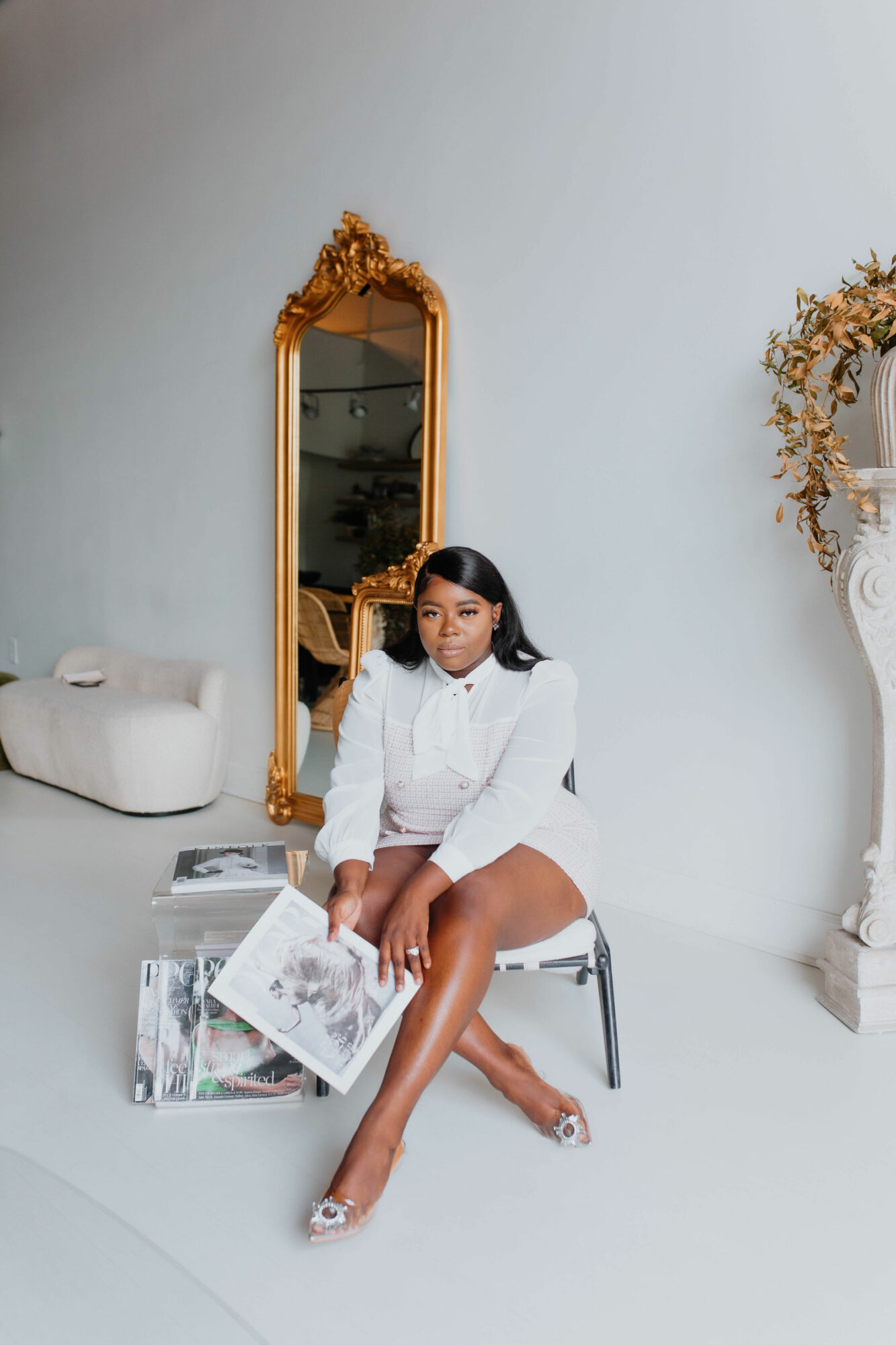 Young woman in white outfit sitting on a chair, holding a magazine, in a room with a large mirror and decorative elements.