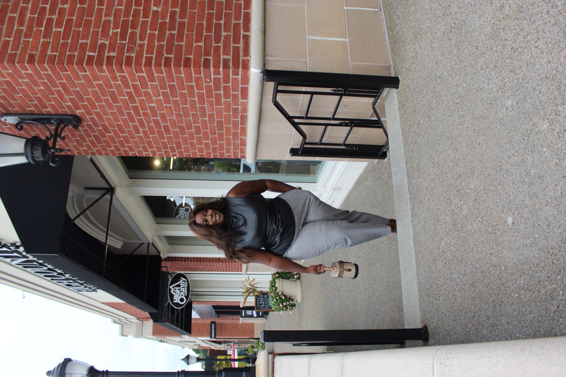 Woman walking on sidewalk outside building with brick and glass facade, plants, and street view in background.