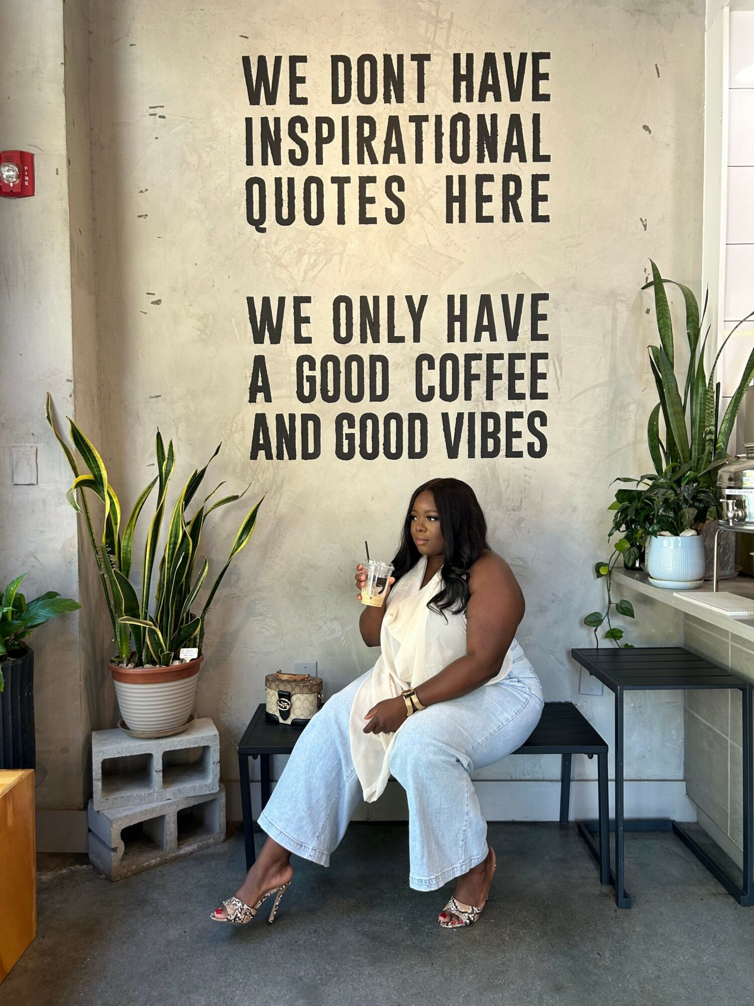 Woman in white dress sitting on a chair, holding a drink, in a modern space with plants and motivational wall quotes.