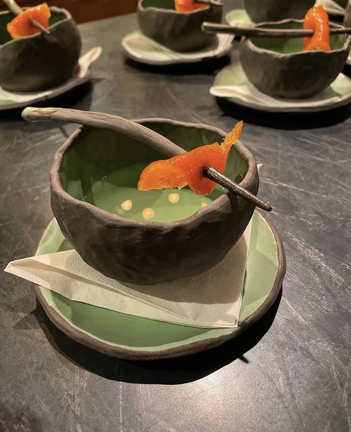 Black bowl with green sauce, orange garnish, and a spoon, on a green plate with a napkin, on a dark surface.
