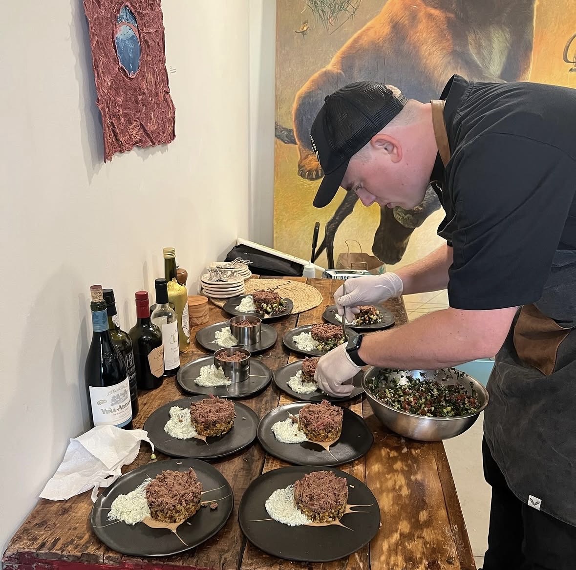 Person preparing plates of food on a wooden table with various dishes, in a room with animal artwork on the wall.