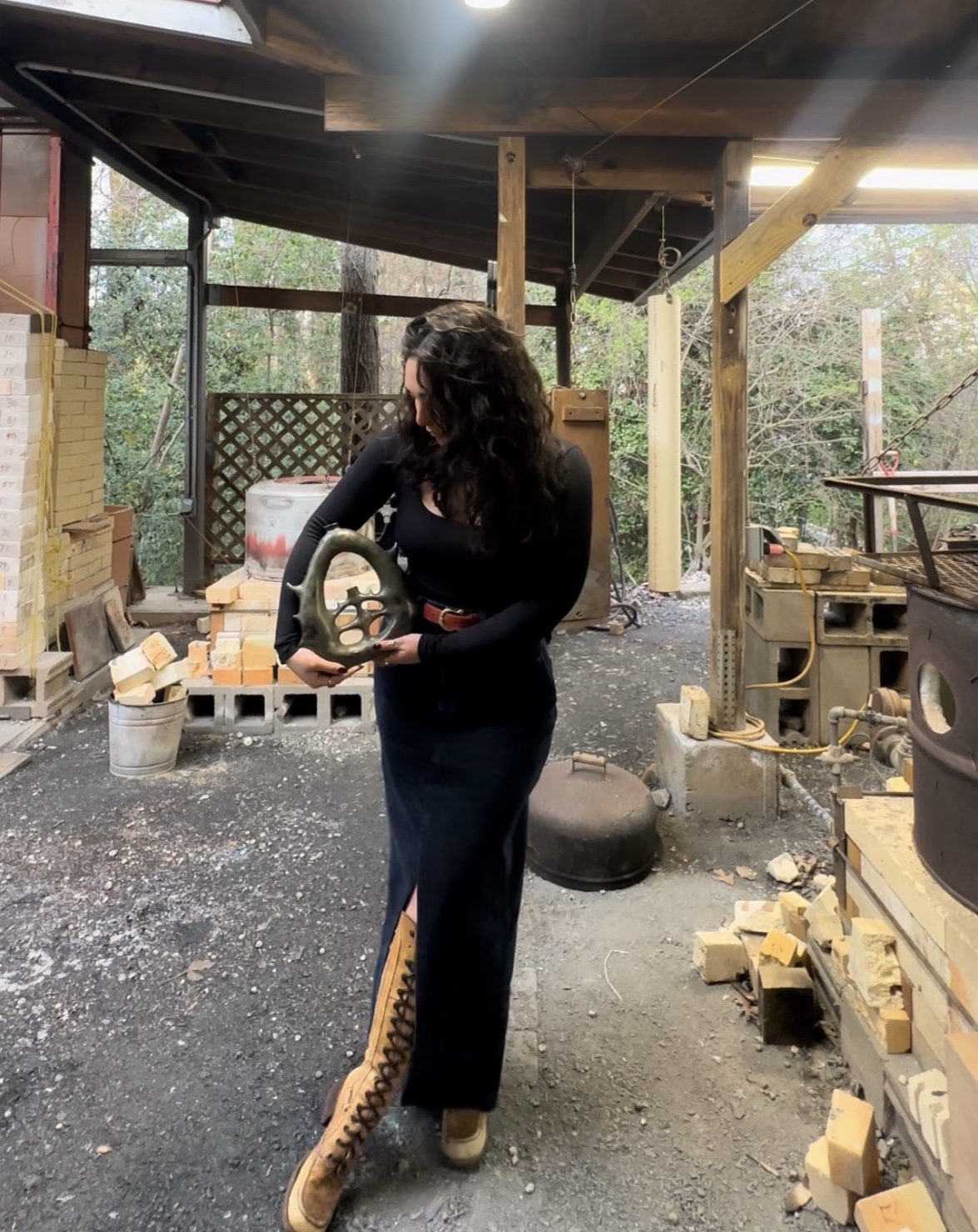 Woman with curly hair holding a circular saw in a workshop with wood and construction materials around.
