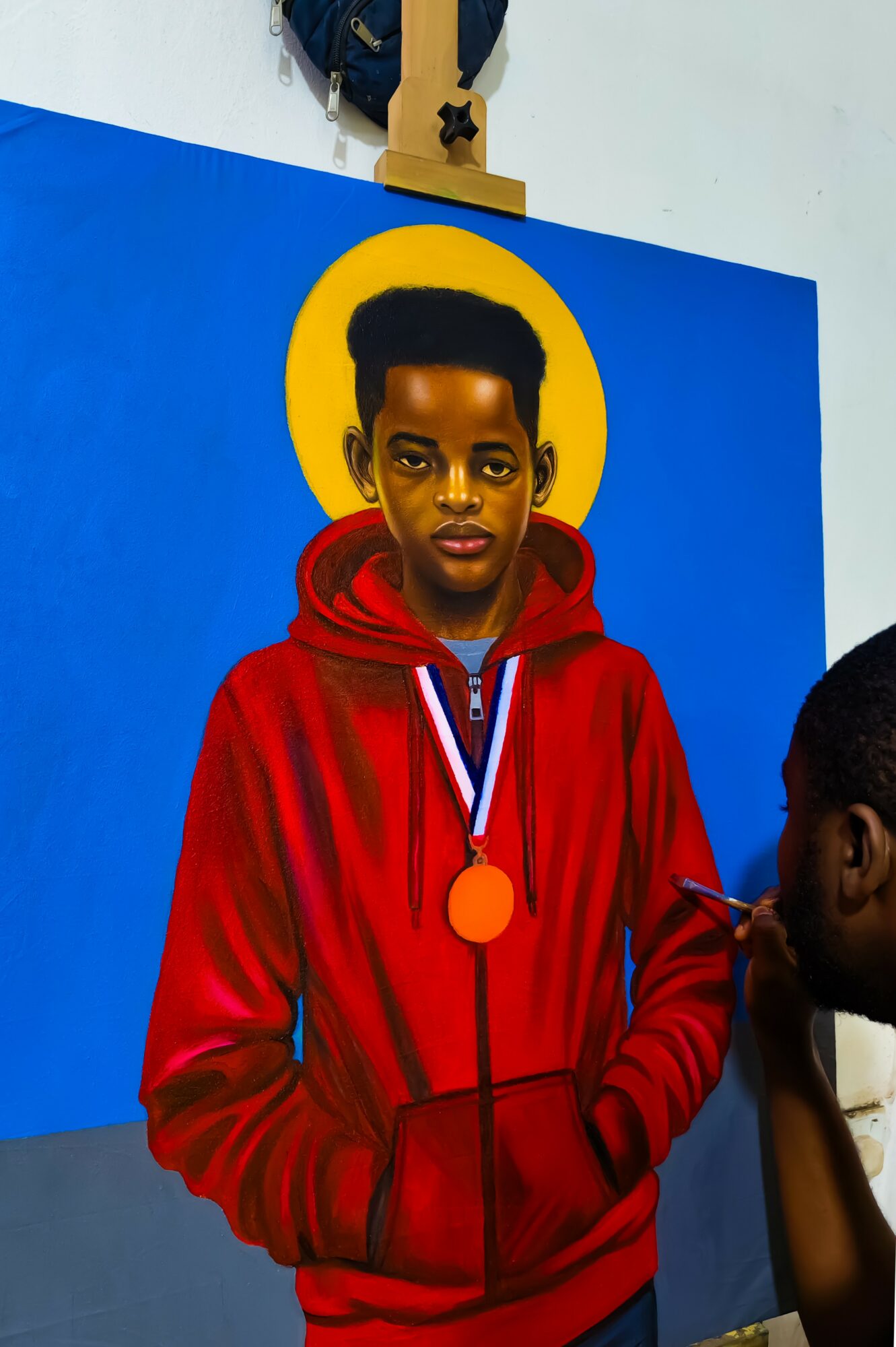 Young person with short hair wearing a red hoodie and a medal, standing in front of a blue background with a yellow circle.