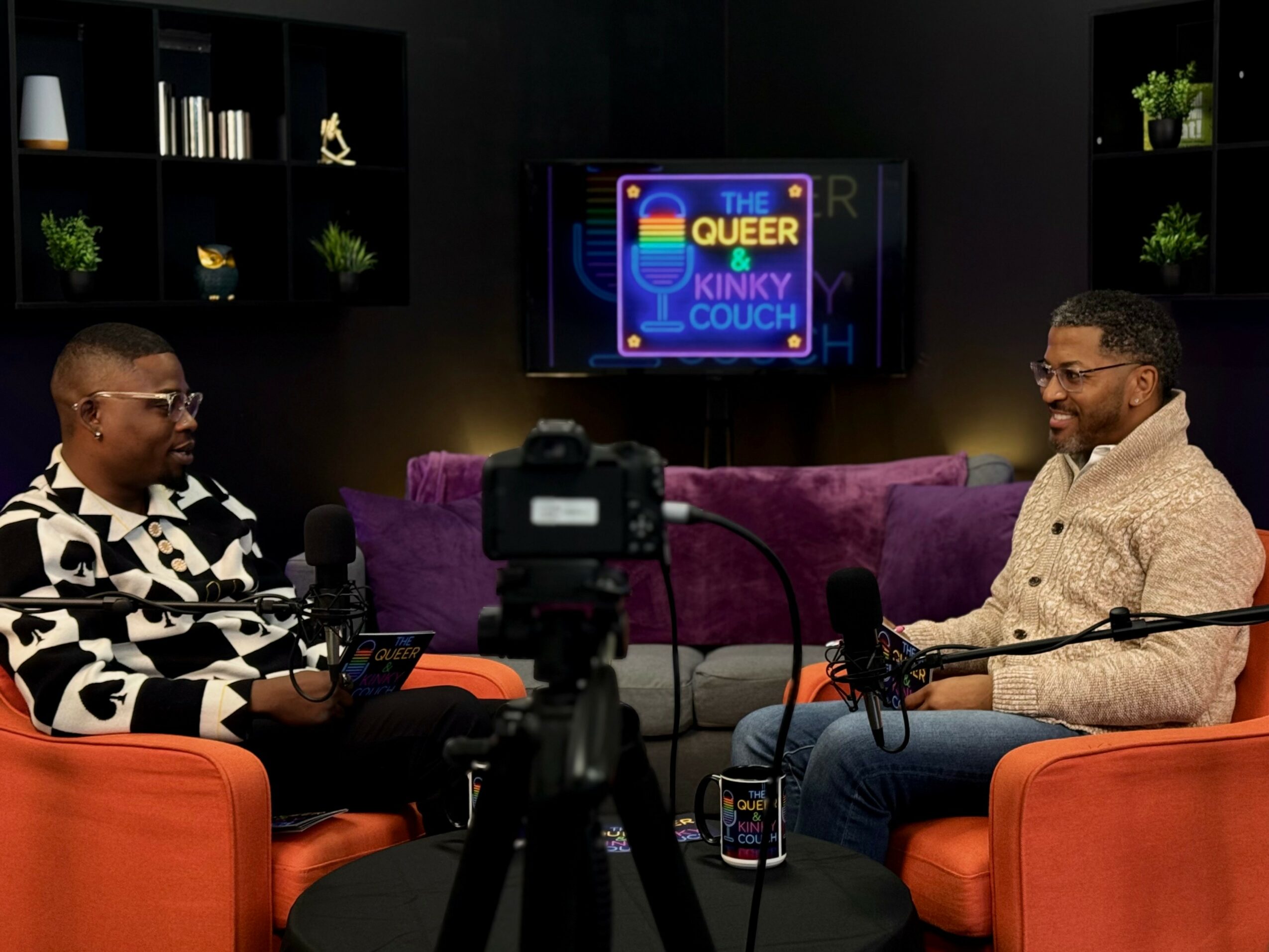 Two men sit on purple and orange chairs in a studio, talking with a camera recording them, and a screen behind them displays a colorful logo.