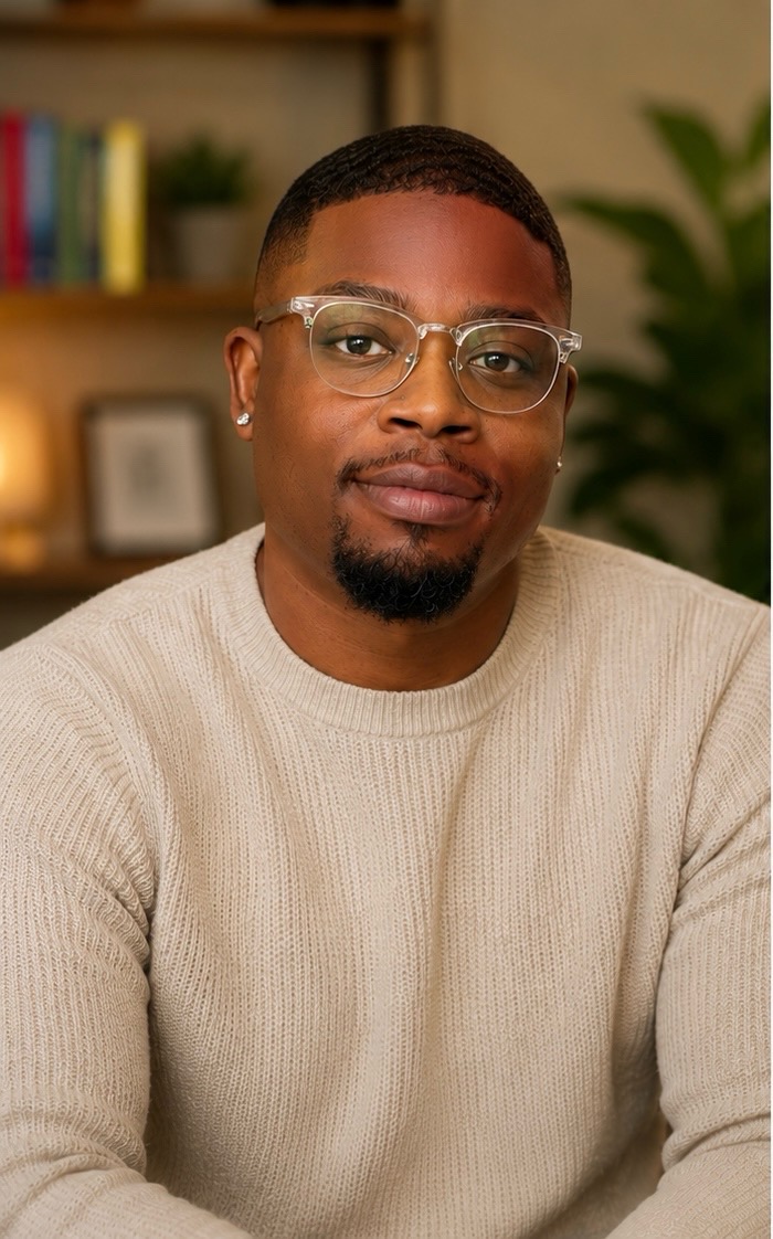 Man with glasses and a beard wearing a beige sweater, sitting indoors with a bookshelf and plant in background.