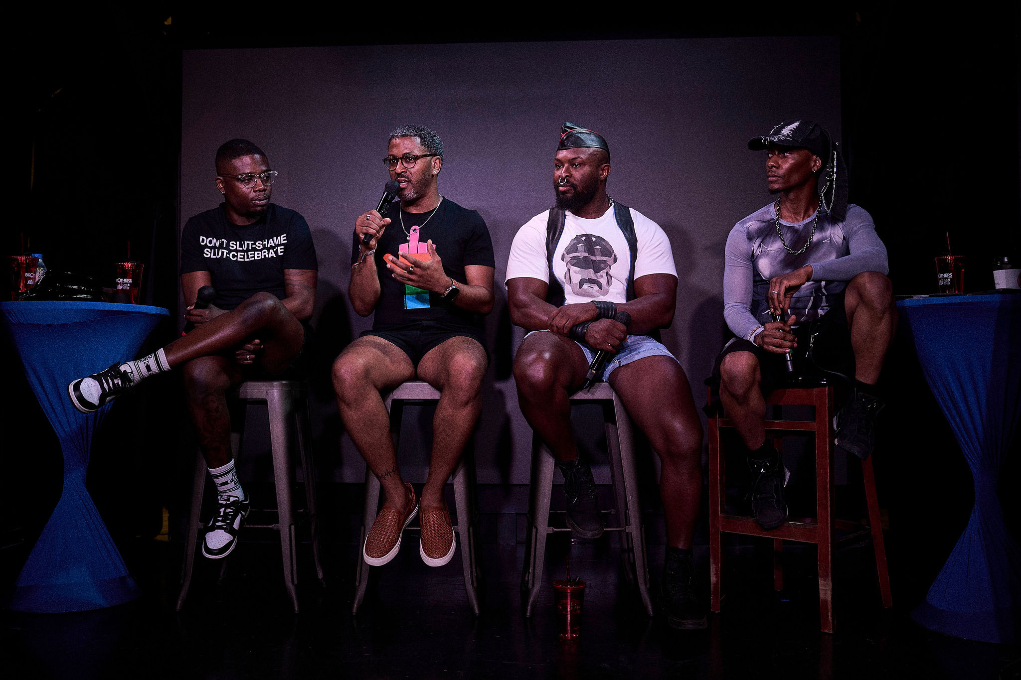 Four people sit on chairs on a stage, engaged in a discussion, with a dark background and blue tables on each side.
