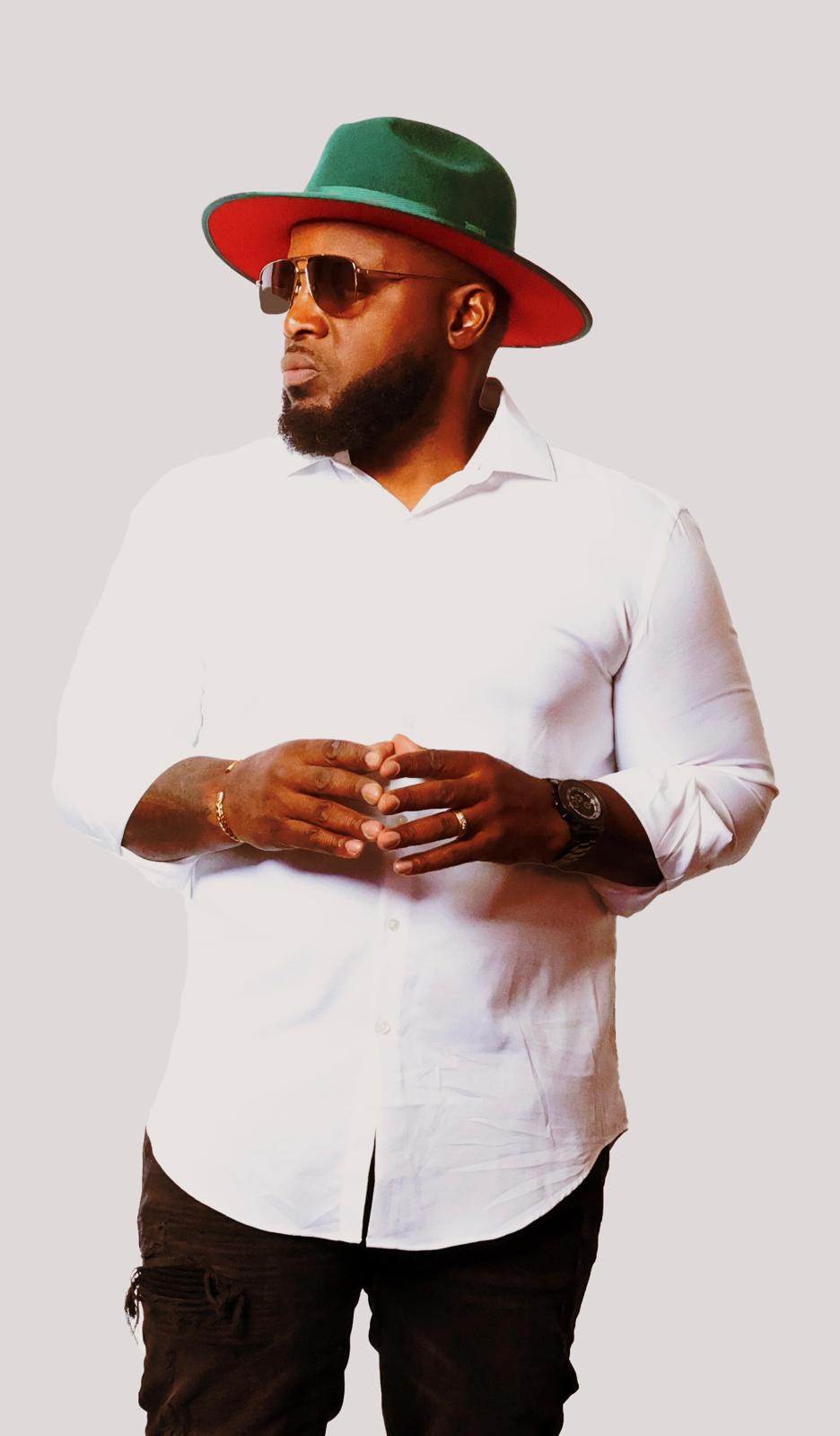 Man with a beard wearing sunglasses, a white shirt, and a wide-brimmed hat with green and red colors, standing with hands clasped.