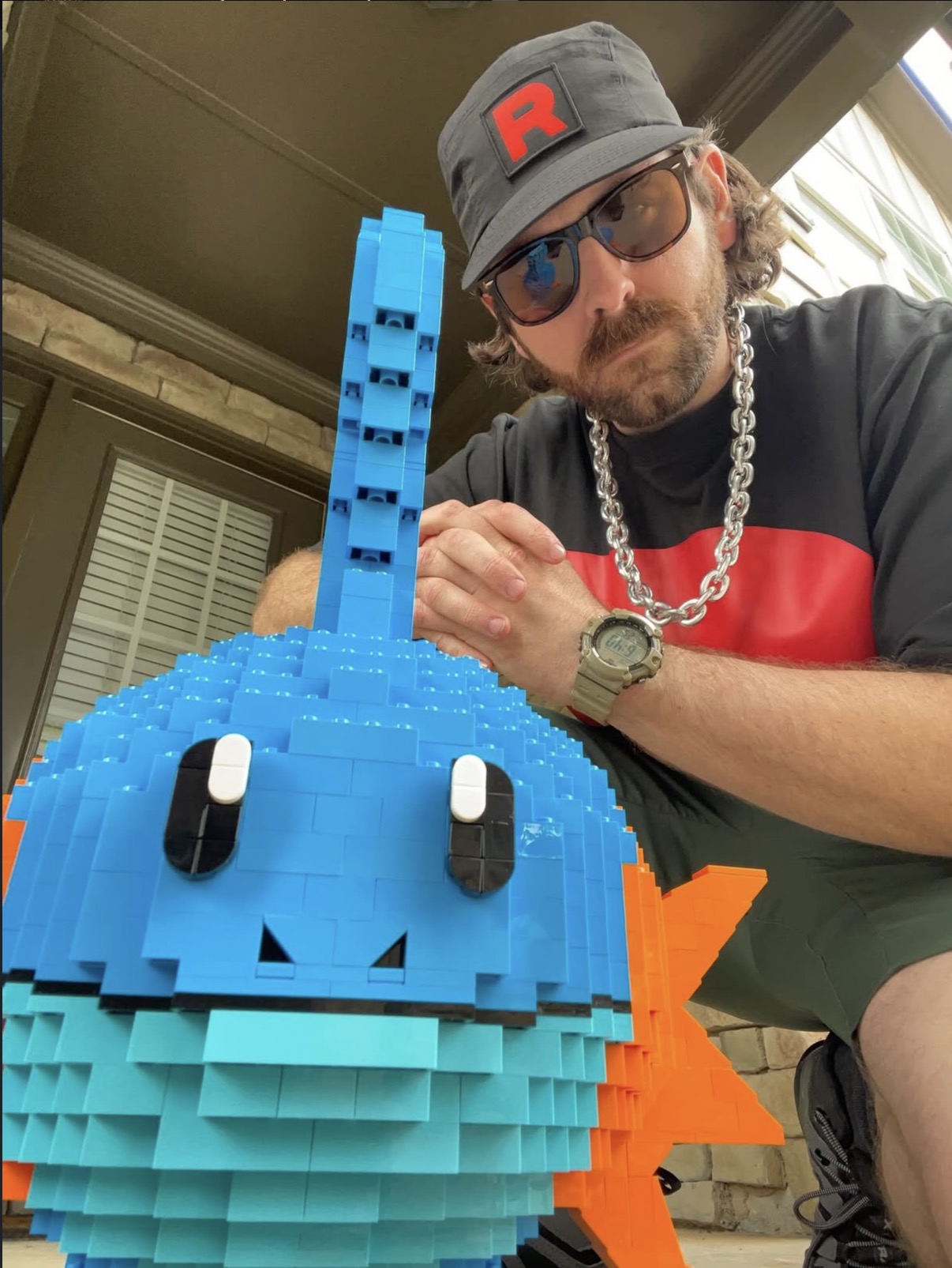 Man wearing sunglasses, a gray cap with a red letter R, and a chain necklace, posing with a large blue LEGO fish sculpture.