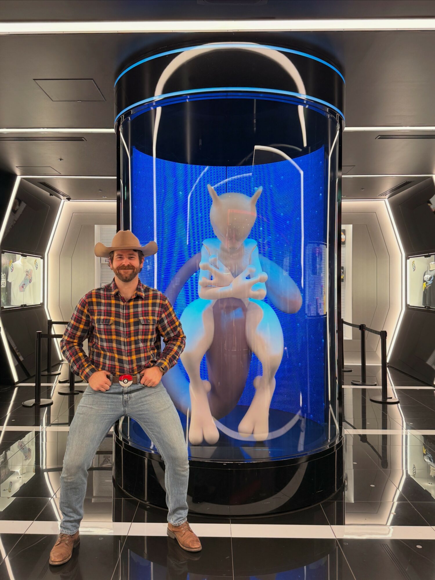 Man in plaid shirt and hat standing in front of a large cylindrical display with a creature inside, in a modern room.