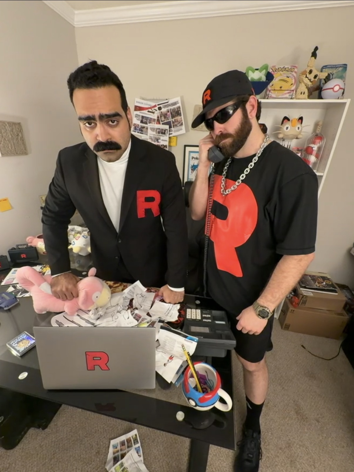 Two men dressed in black with red 'R' logos stand behind a cluttered desk with a laptop, toys, and papers.