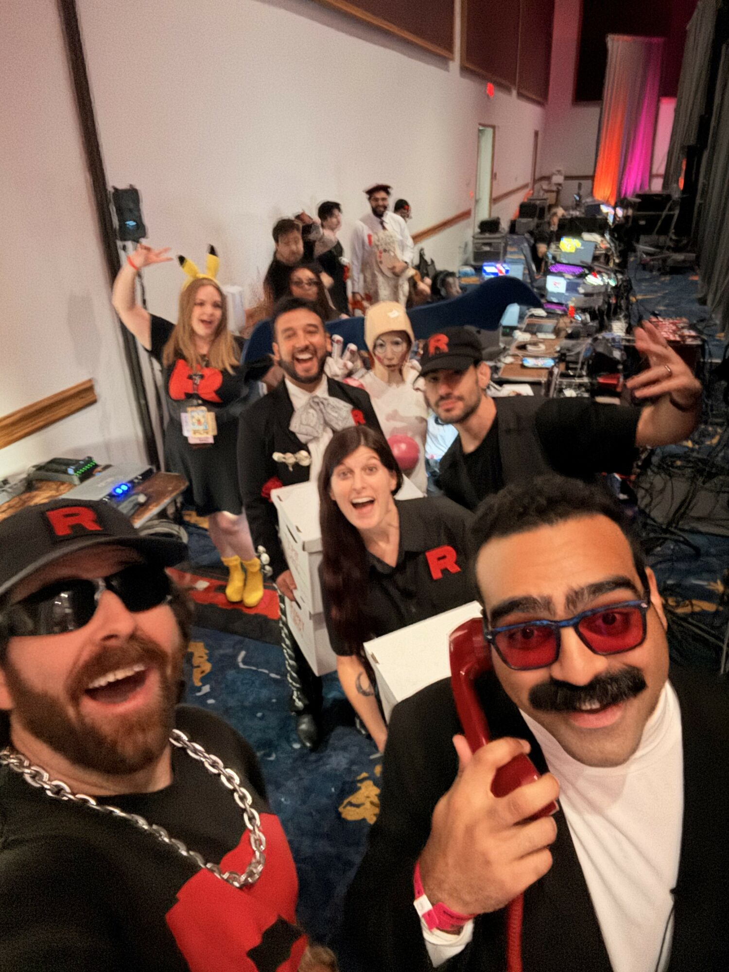 Group of people at a party or event, smiling and posing for a selfie, with a row of tables and equipment in the background.