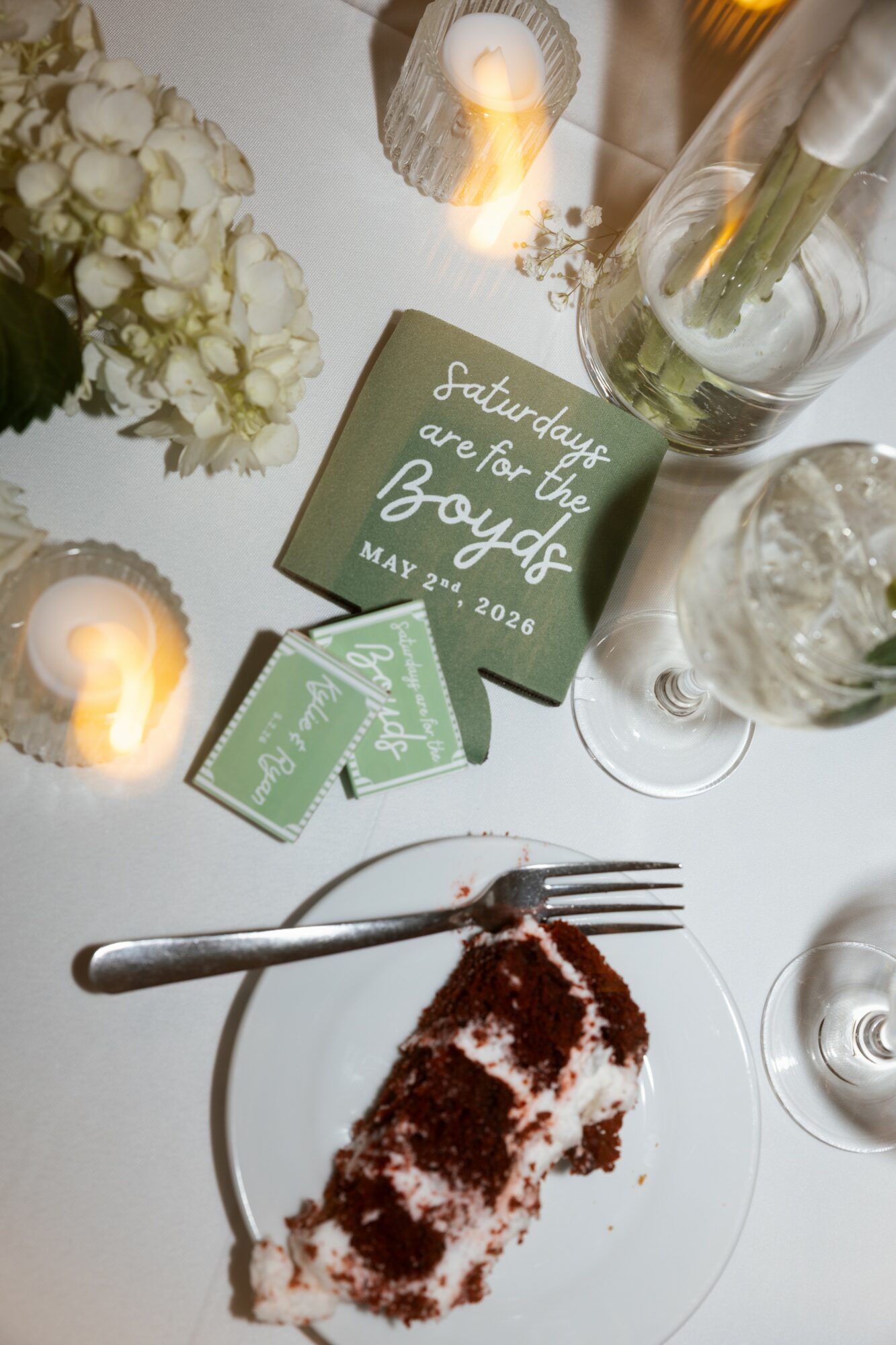 Wedding cake slice on plate with fork, candles, flowers, and a card reading 'Saturday, 2nd of the Boyds, May 23, 2020' on table.