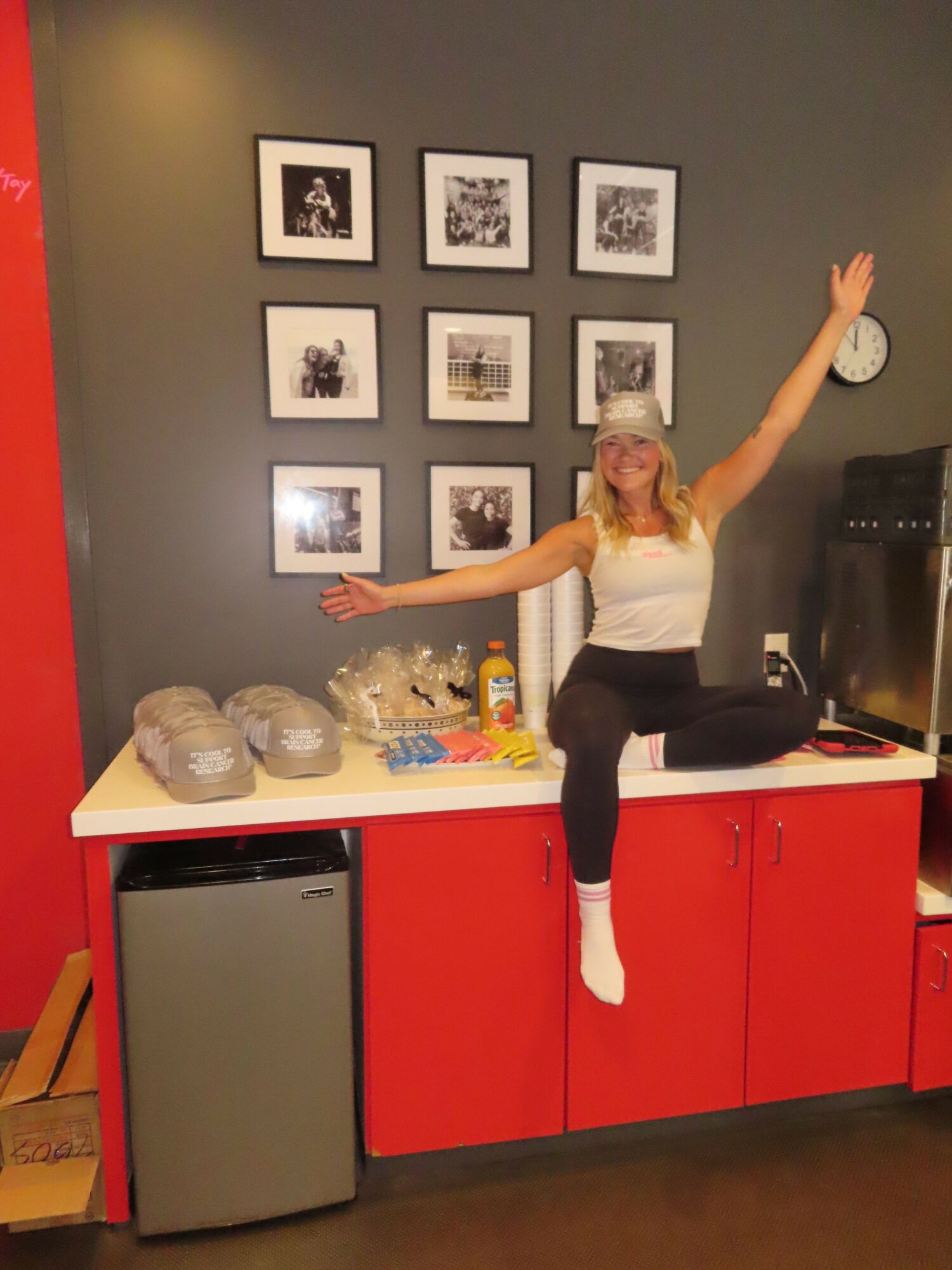Person standing on one leg with arms outstretched in a room with framed pictures, red cabinets, and a table with items.