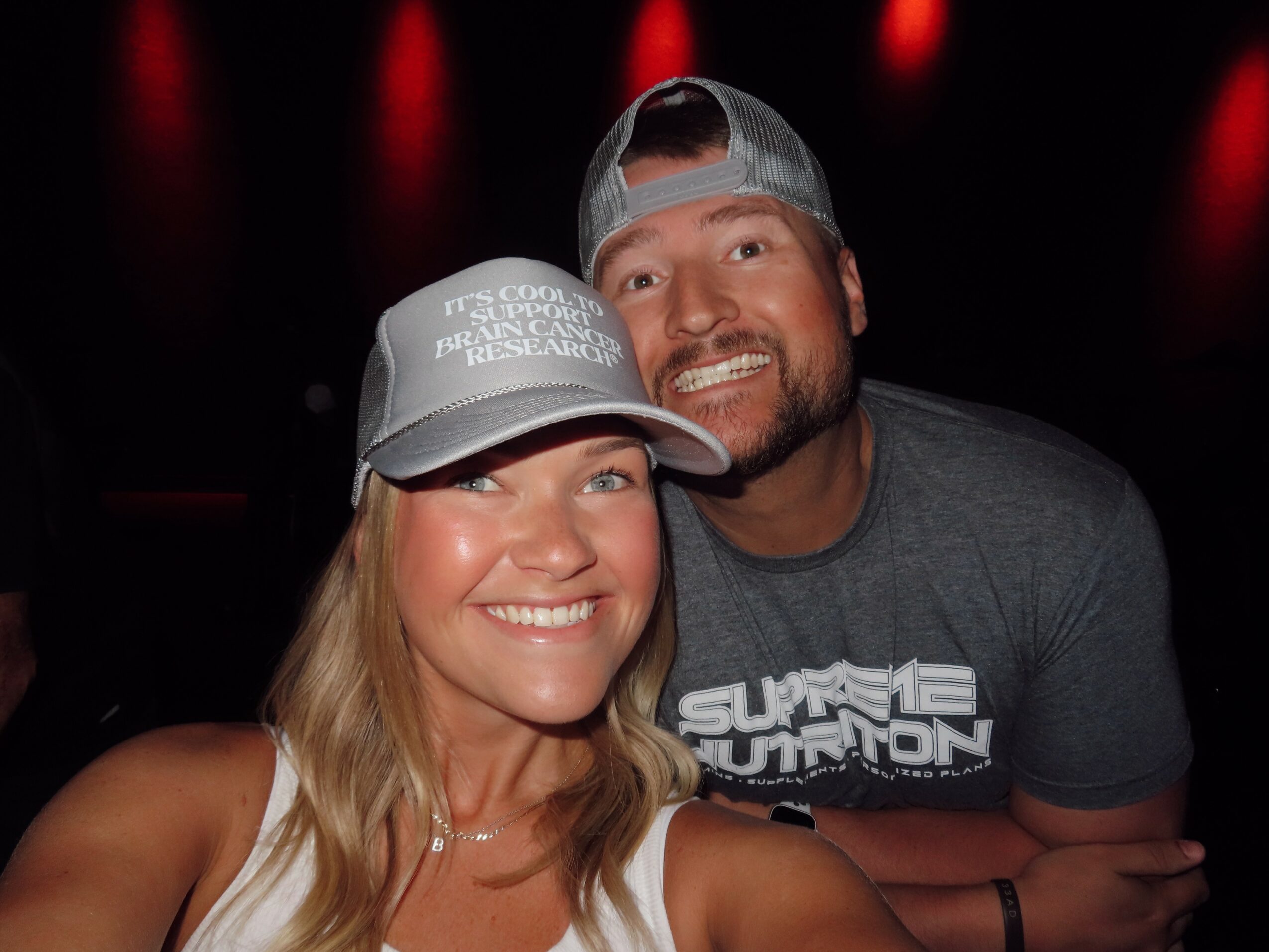 Two smiling people, a woman and a man, wearing caps, close together in a dark setting with red lights.