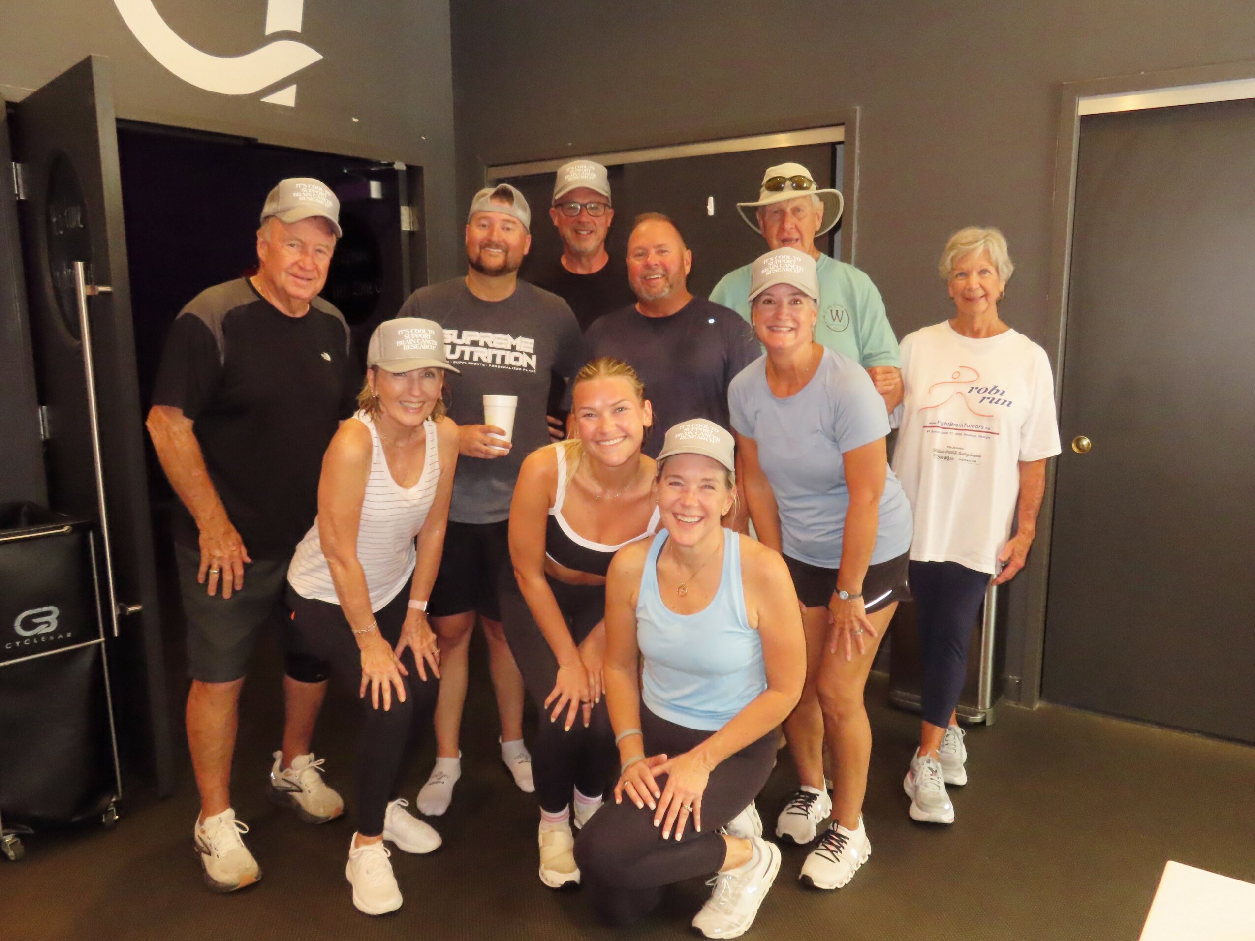 Group of ten people smiling indoors, some wearing hats and casual clothing, standing and kneeling together.