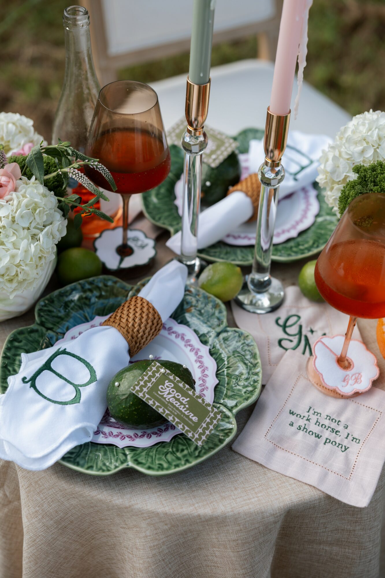 Table setting with plates, glasses, candles, flowers, and a napkin with a green tag, outdoors with greenery in background.