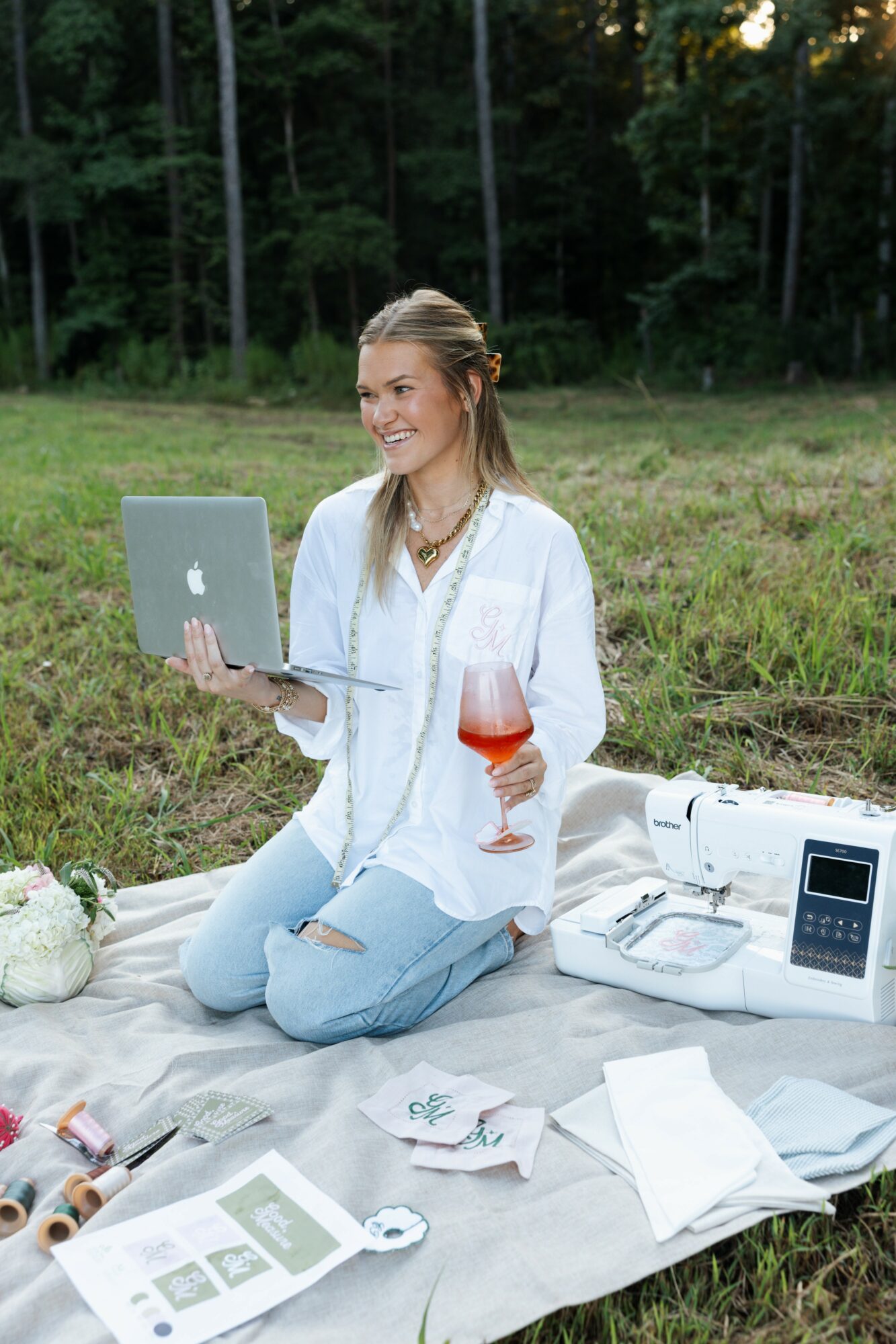 Woman sitting on a blanket outdoors with a laptop, drink, and various items, smiling and looking to the side.