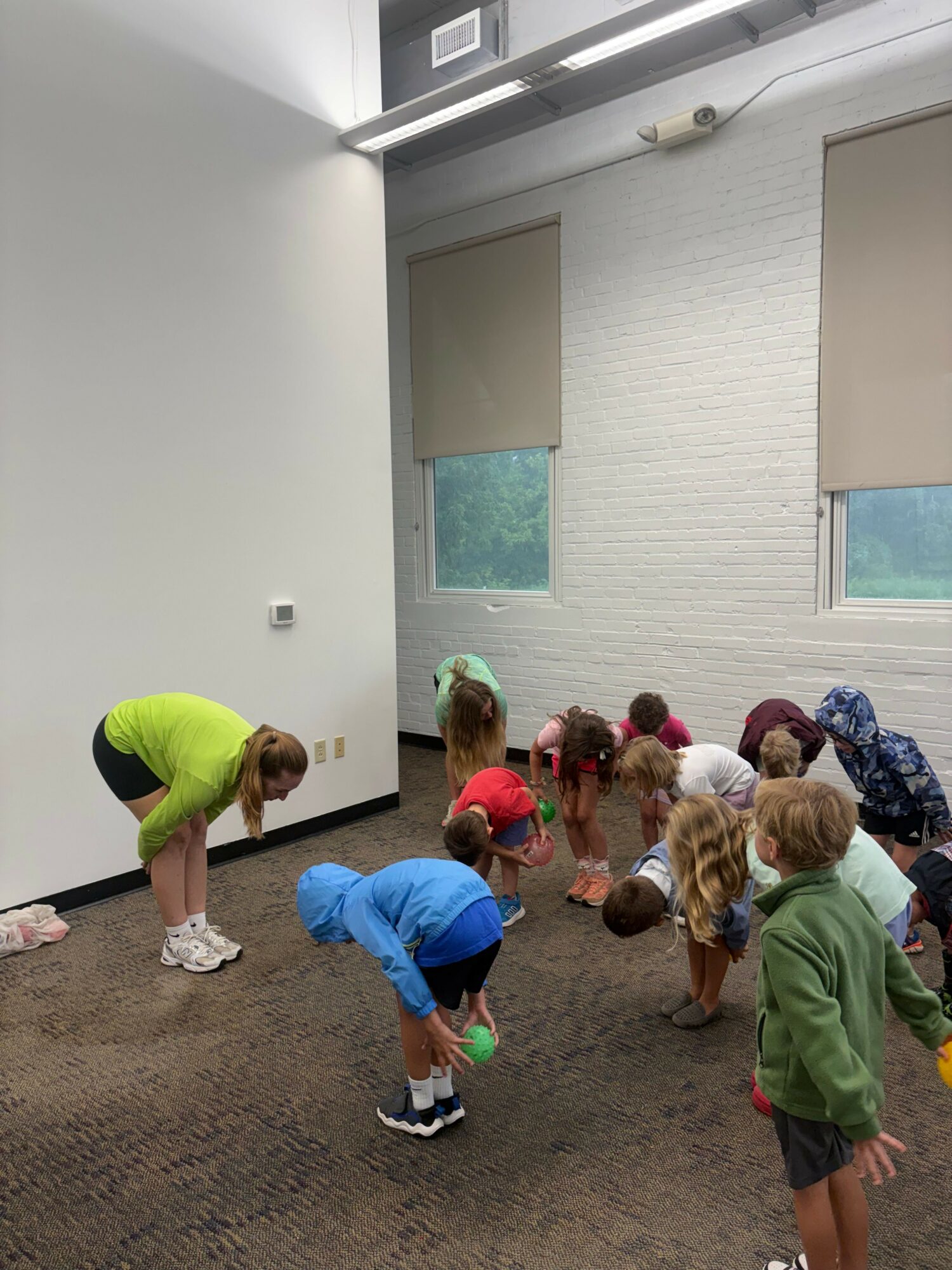 Group of children gathered around a person kneeling on the floor in a room with white walls and windows.