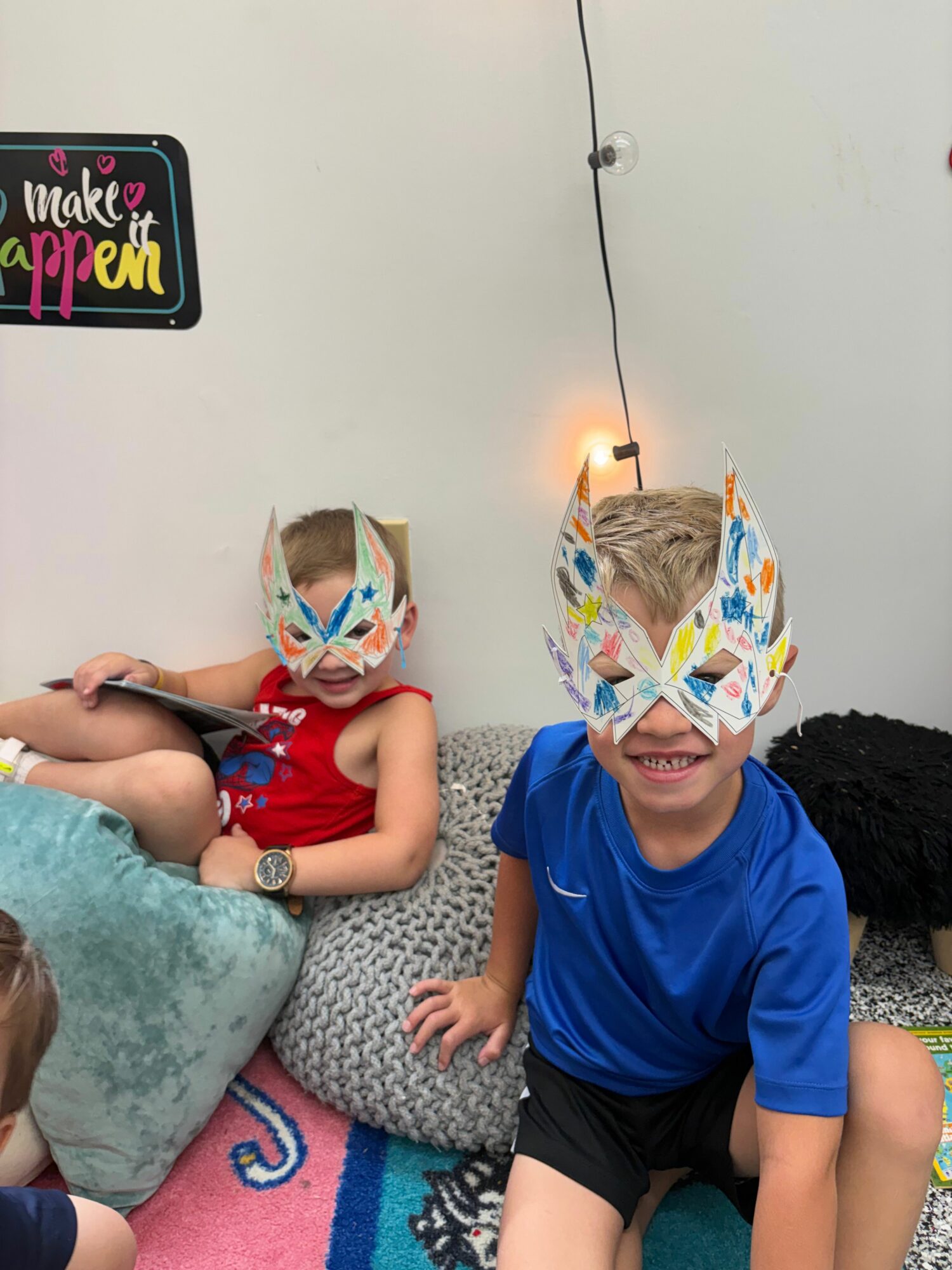 Two children sit on a colorful rug, wearing paper hats, smiling, with a gray pillow and a wall with a light fixture behind them.