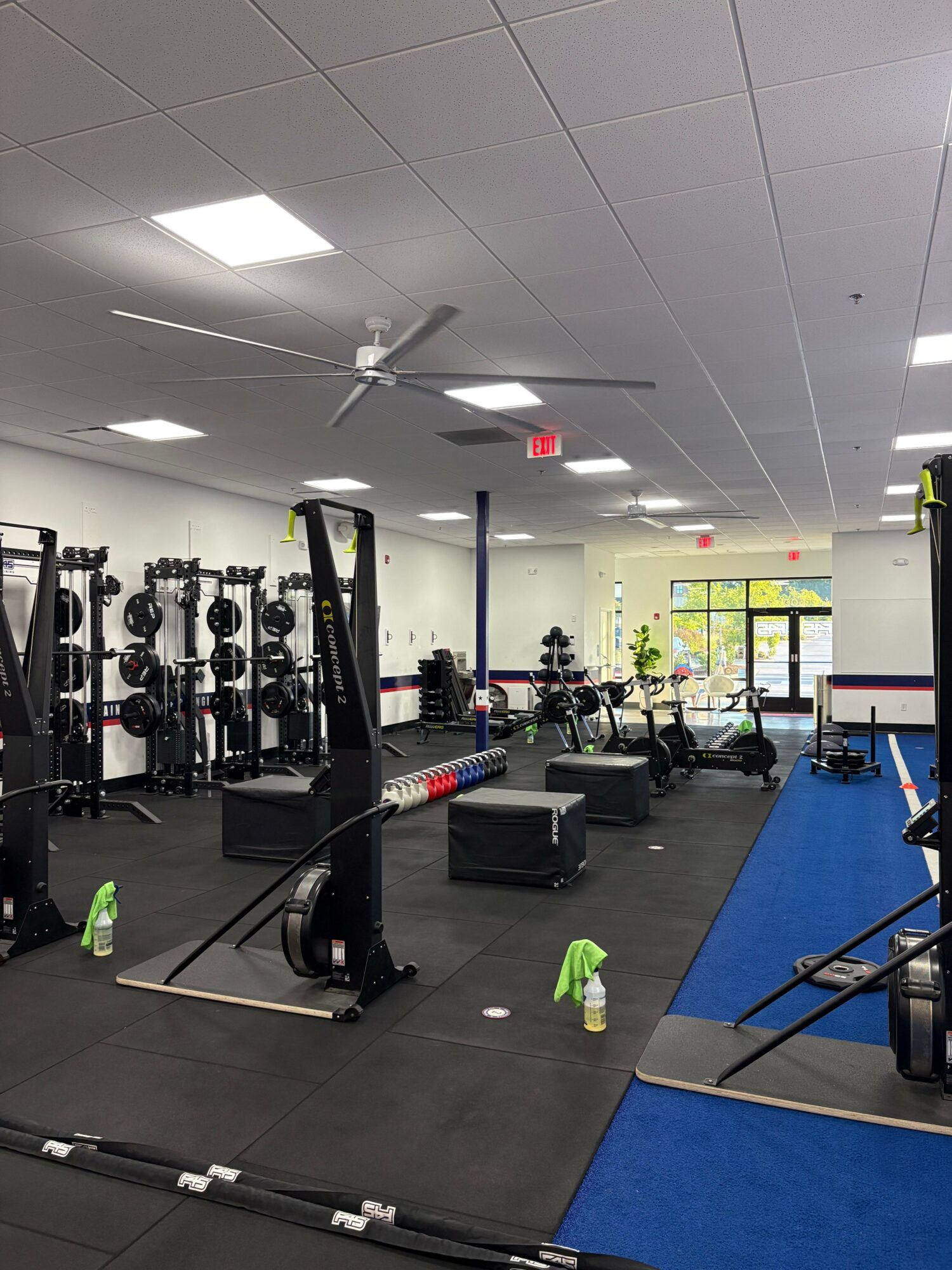 Gym with workout equipment, mats, and people exercising, viewed from the entrance towards the back of the room.