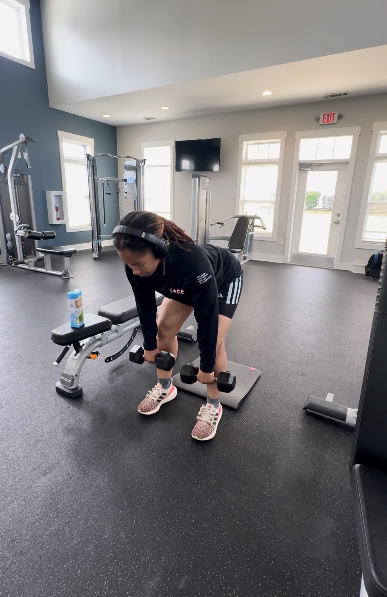 Woman in black workout clothes bending over in a gym, holding dumbbells, with exercise equipment and windows in the background.