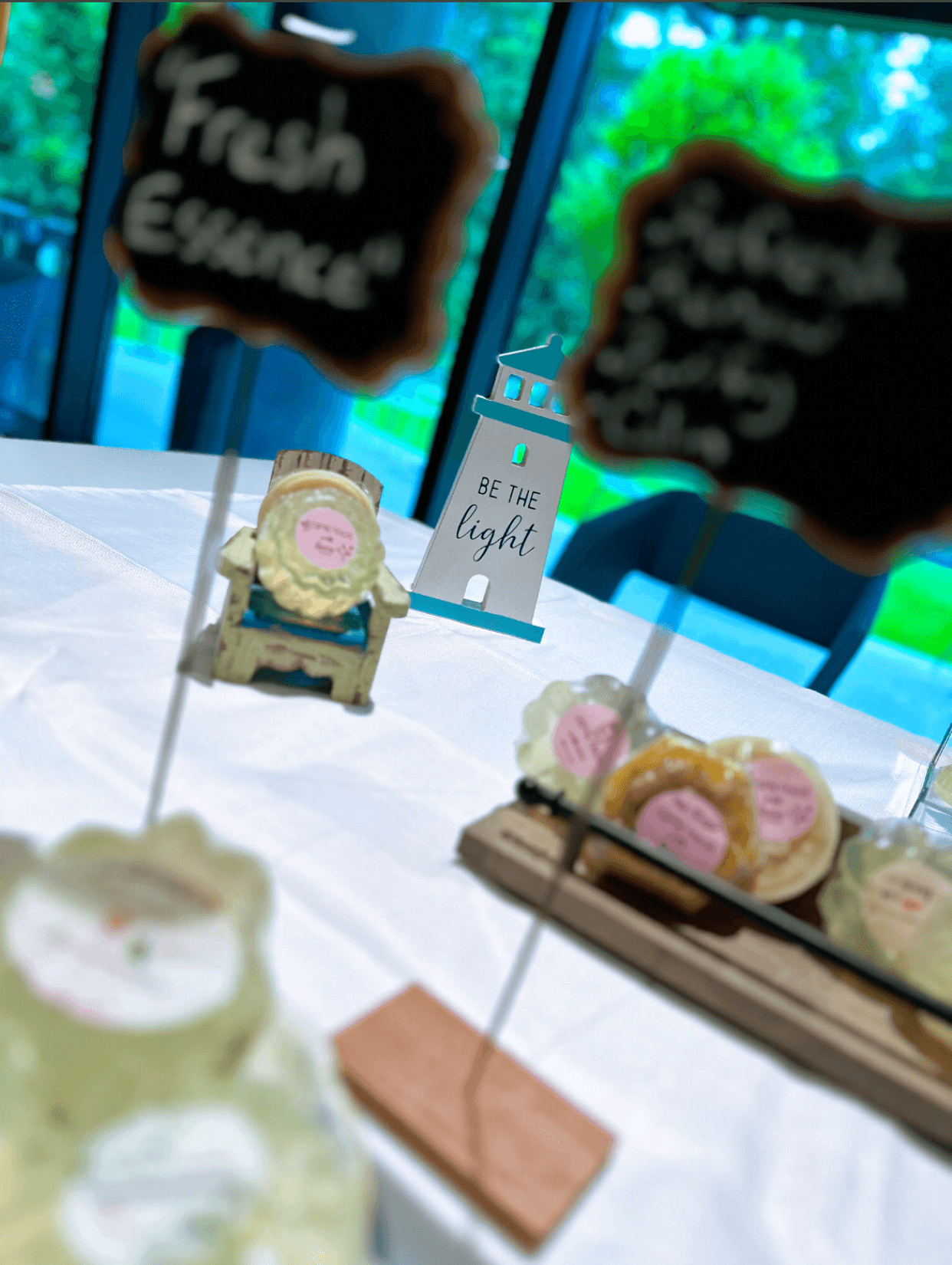 Table with baked goods, chalkboard signs, and a small sign reading 'Be the light' in front of a window with trees outside.