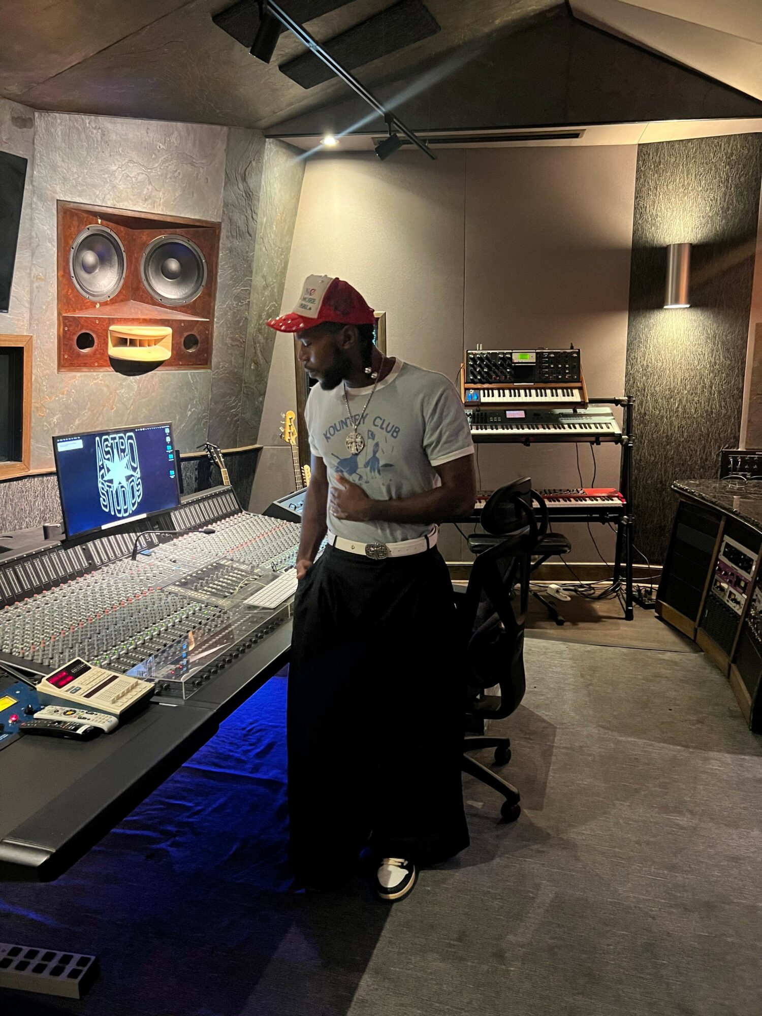 Person wearing a red cap and white shirt stands in a music studio with audio equipment and speakers.