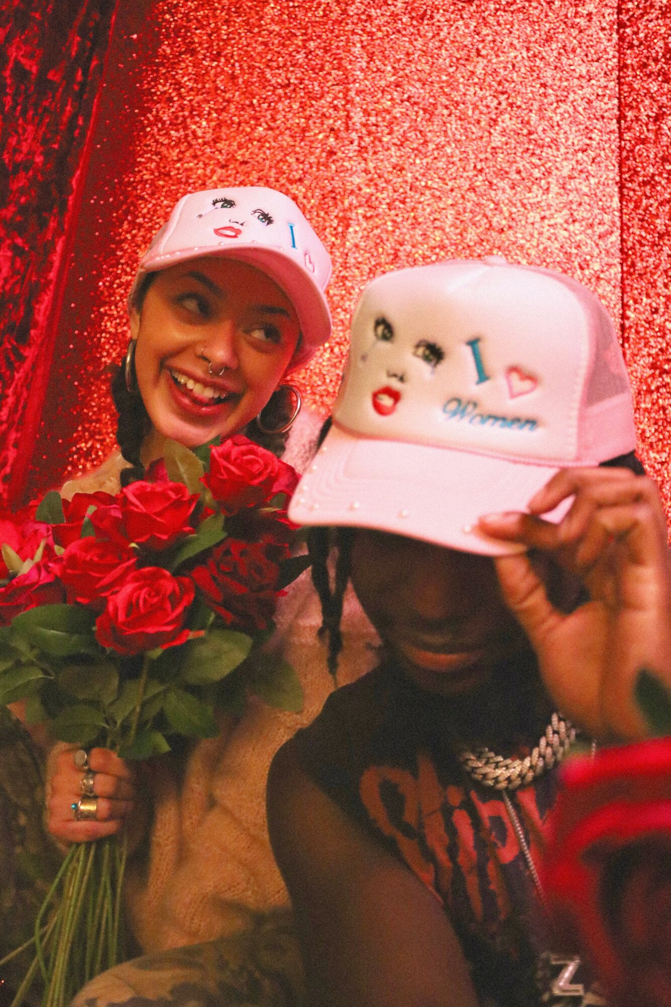 Two people wearing white hats with cartoon faces, one holding a bouquet of red roses, against a red background.
