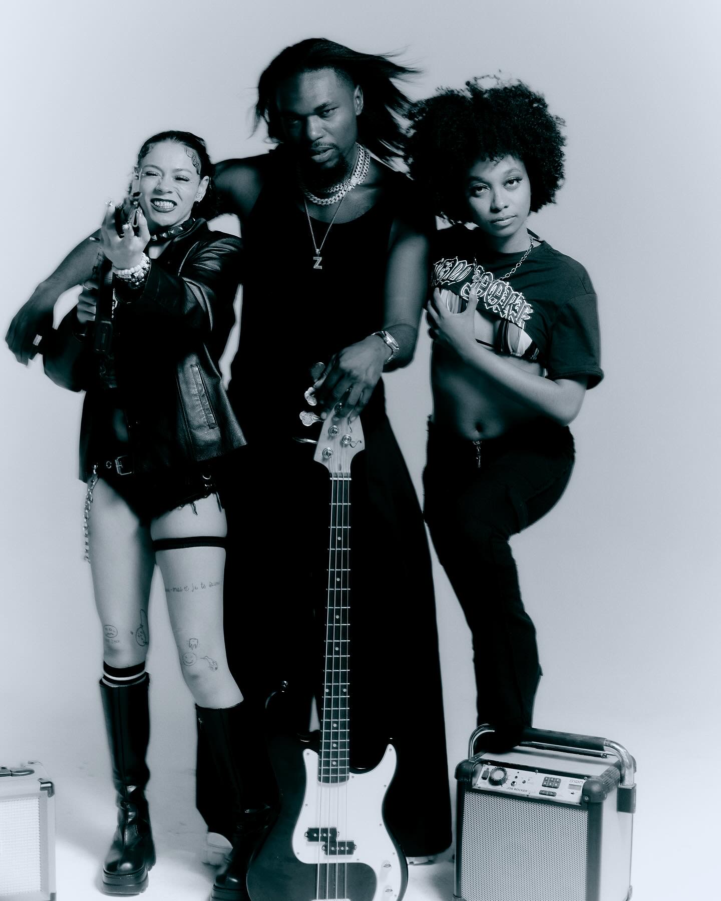 Three people posing together, one holding a guitar, with a small amplifier nearby, against a plain background.