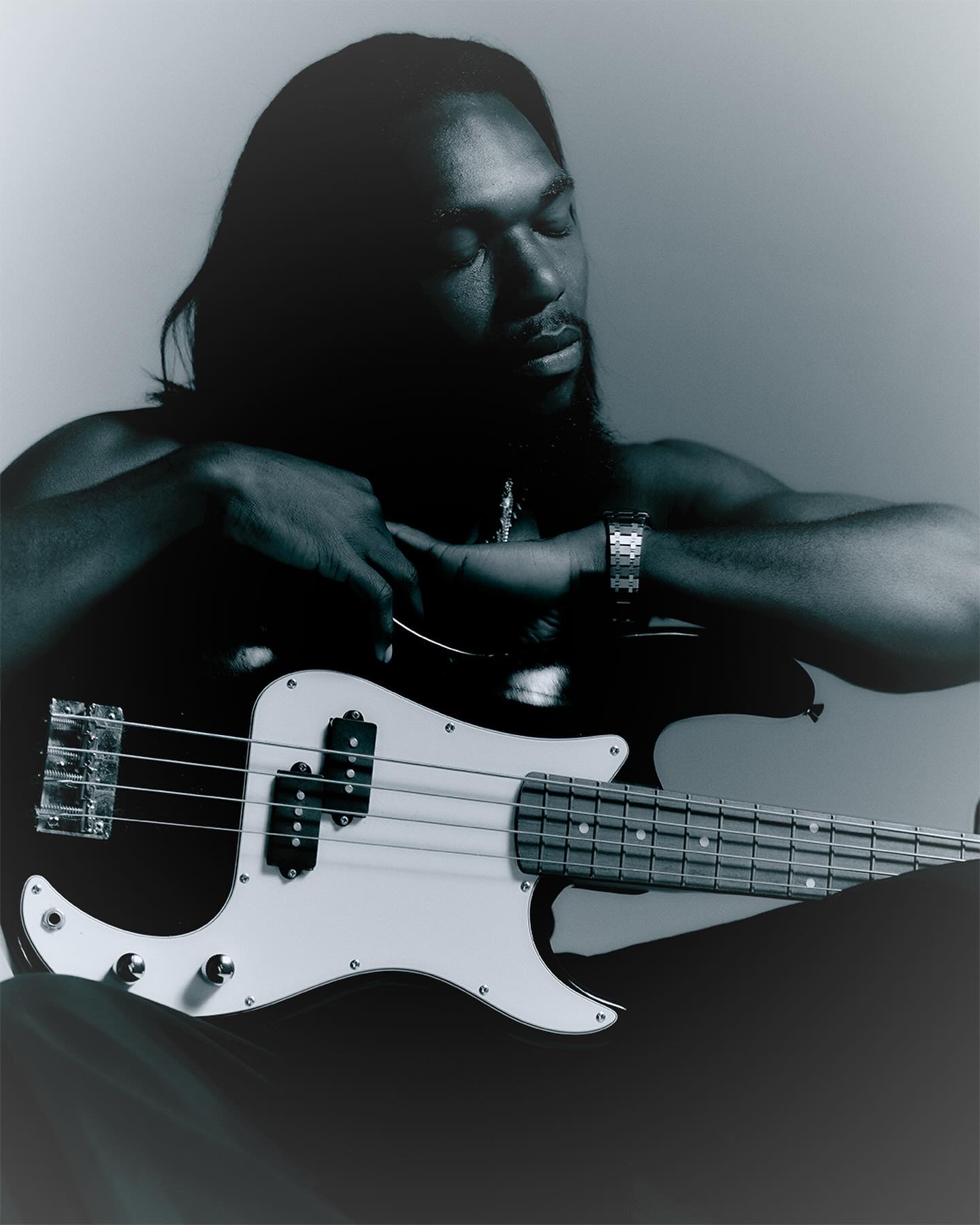 Person with long hair holding a bass guitar, eyes closed, in a dark setting.