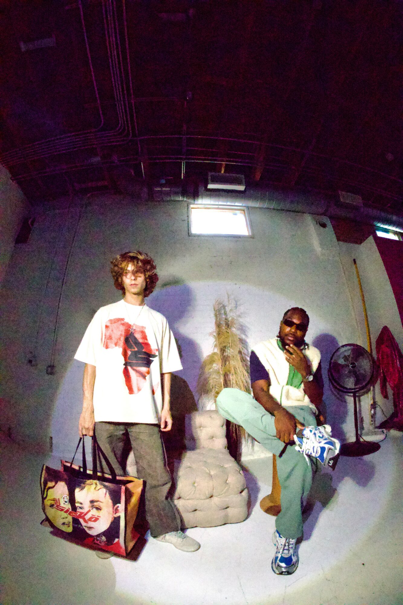 Two people in a room with a large bag, a sculpture, and fans, one sitting and one standing, with a window above.