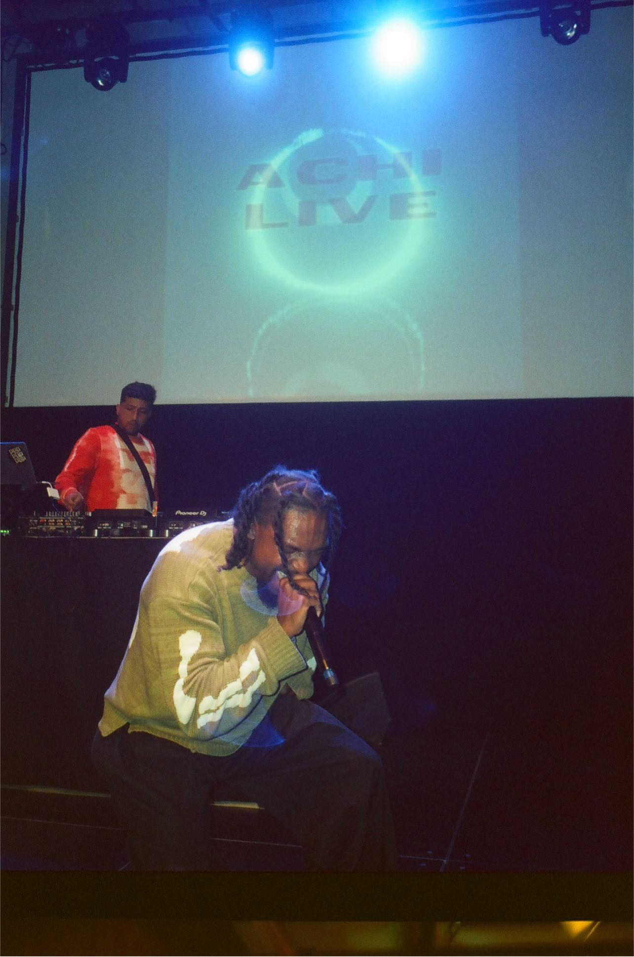Person with glasses and a white shirt sitting on a stage, with a DJ in the background and a projected logo on the screen behind.