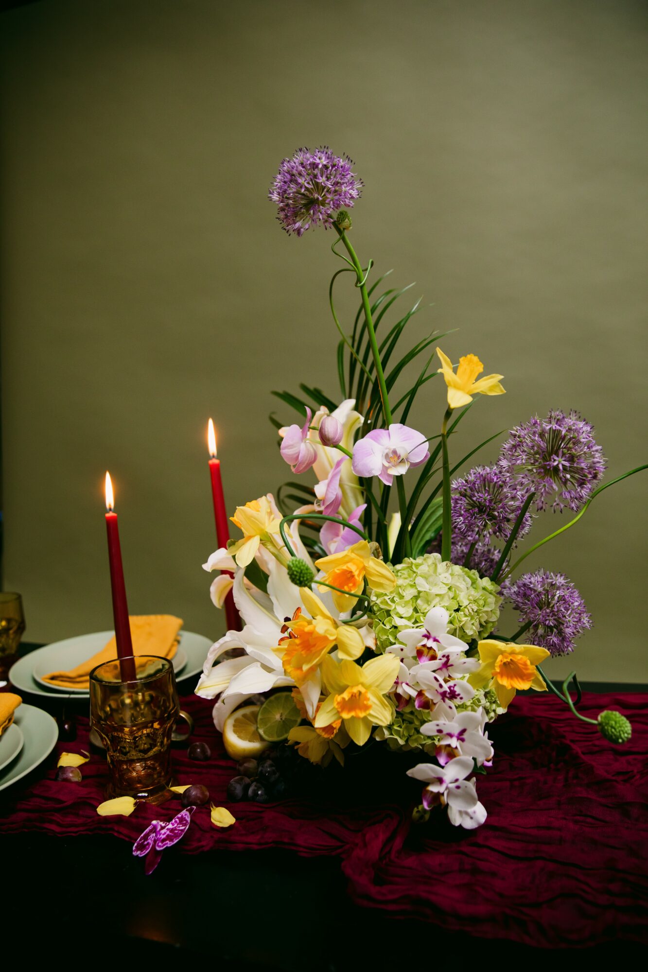 Flower arrangement with purple, yellow, and white flowers, three red candles, and a dark tablecloth.