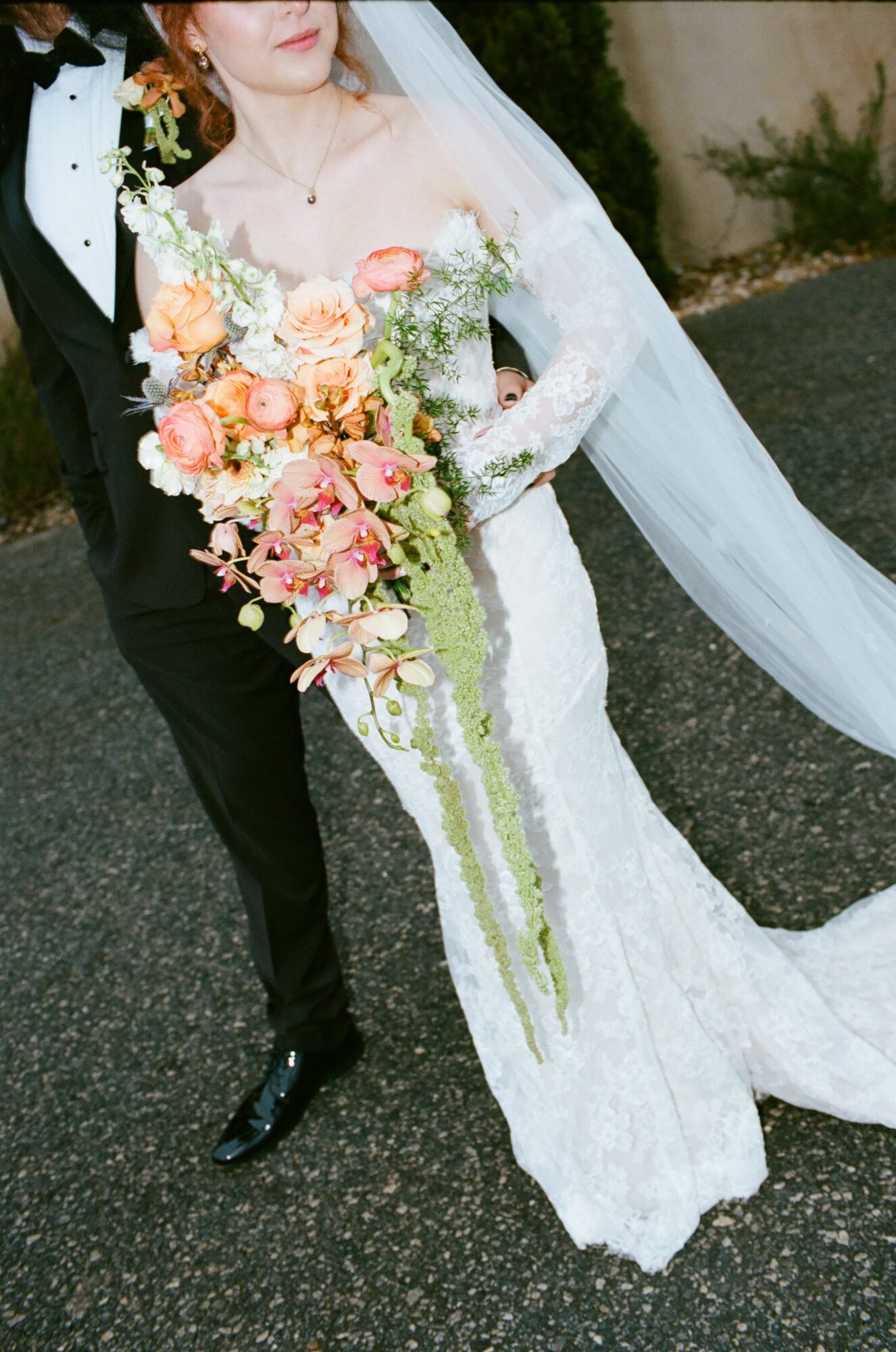 Bride in a white lace wedding dress holding a large bouquet of pink, peach, and white flowers, with a veil flowing.